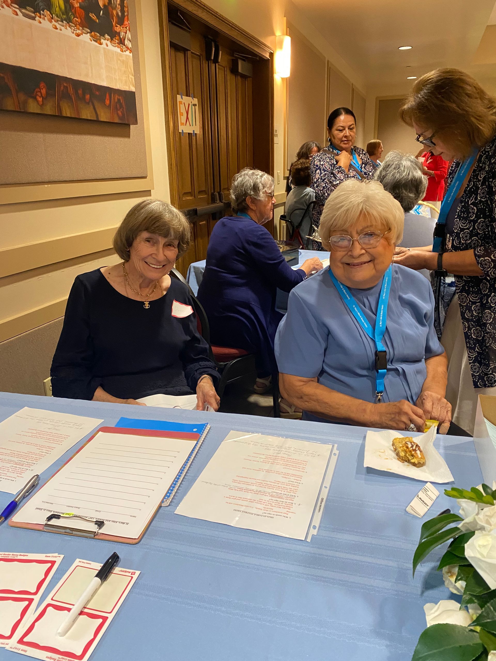 Two women smiling at a registration table with forms, pens, and a blue tablecloth in a hall