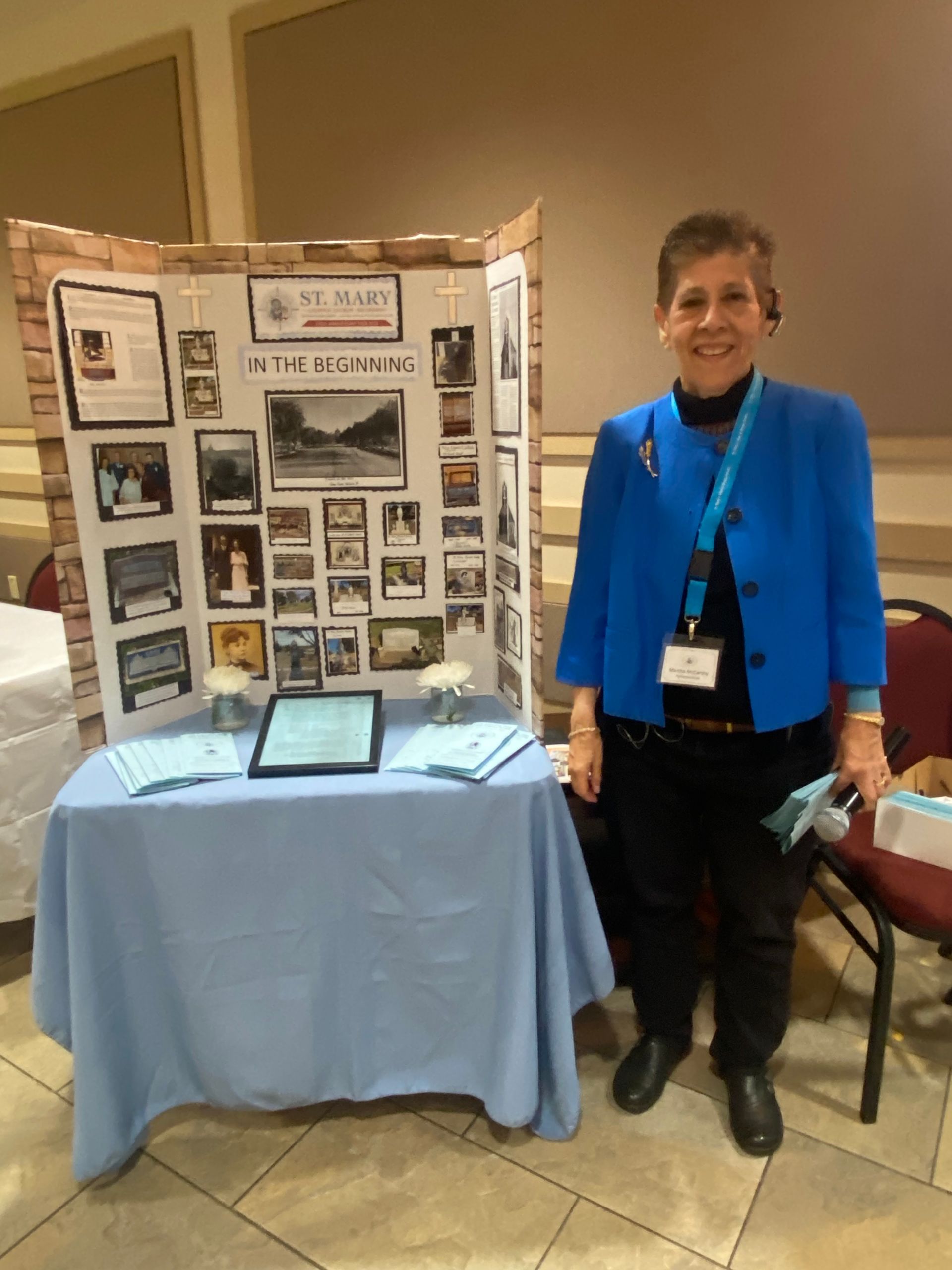Woman stands beside a display booth with photos, a tablet, and a light blue tablecloth.