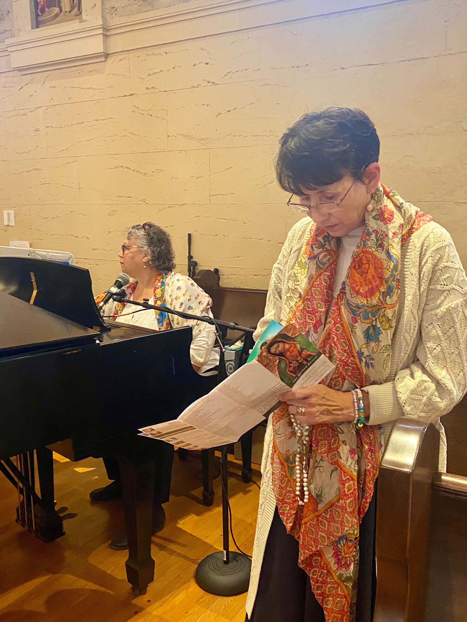 Person reading from a sheet beside a grand piano, with another seated at the piano in a hall.