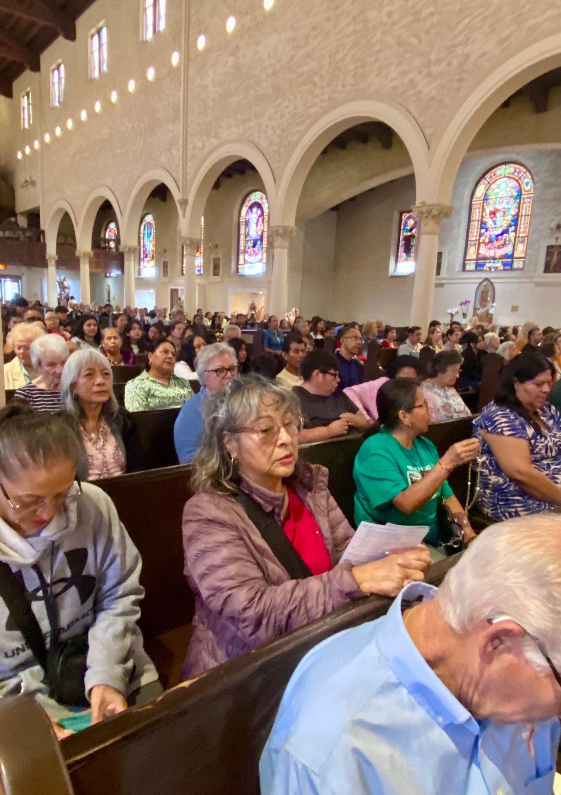 Crowded church service with people seated in wooden pews beneath arched ceilings and stained glass windows