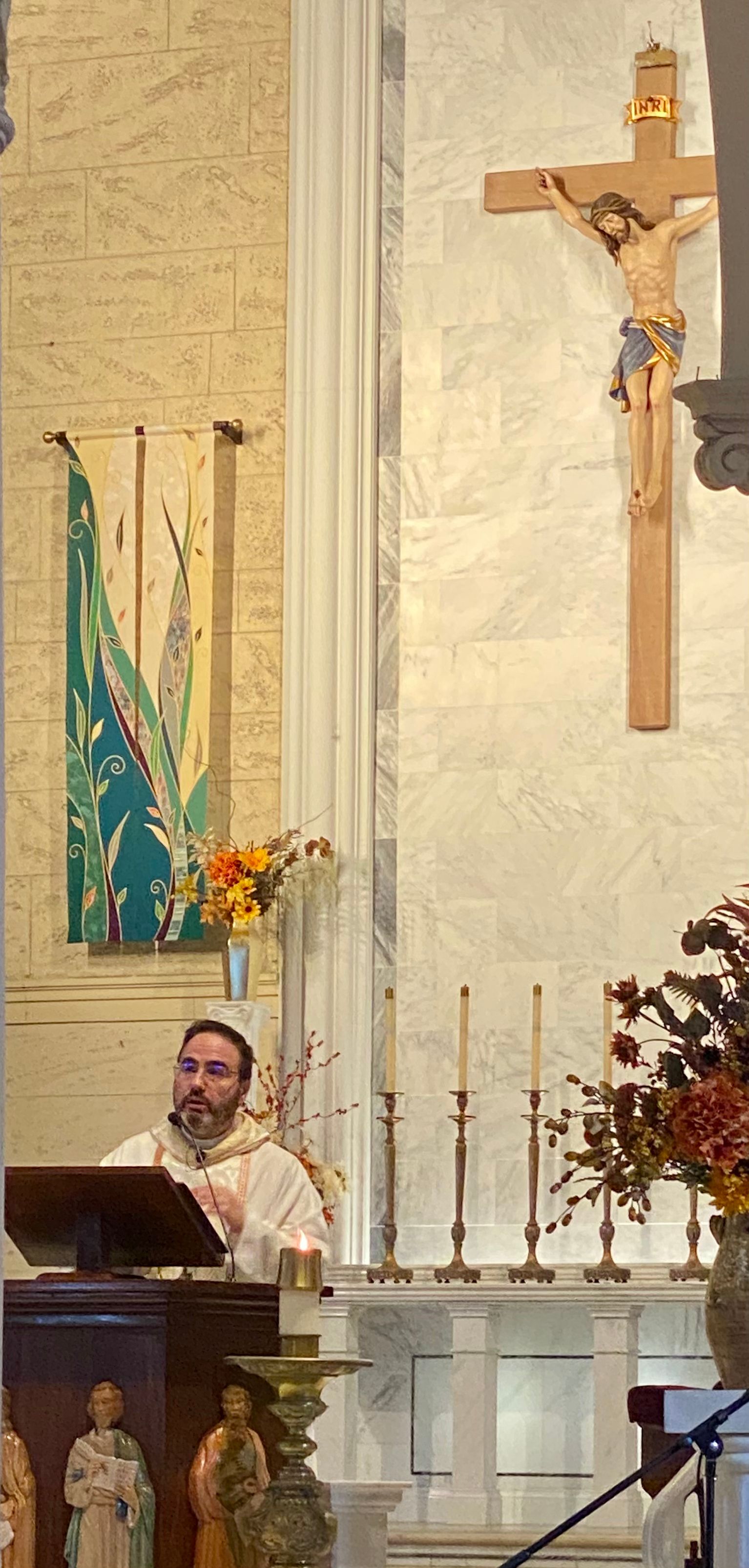 Priest speaking at a church lectern beside candles and a crucifix altar backdrop