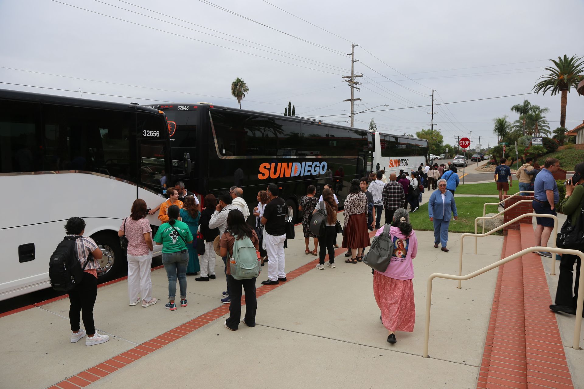 People waiting to board a white and black tour bus at a bus terminal on a cloudy day