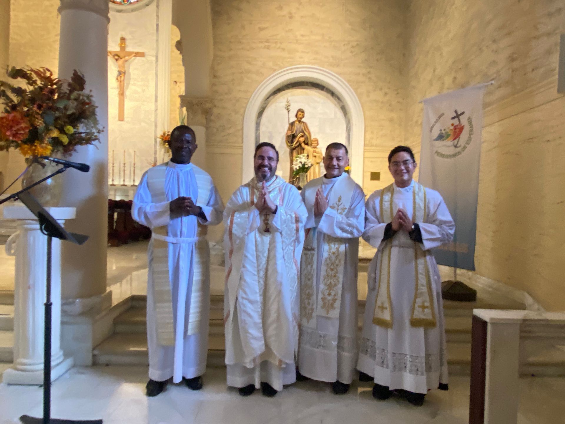 Four clergy in white robes stand in a church before an altar and crucifix.