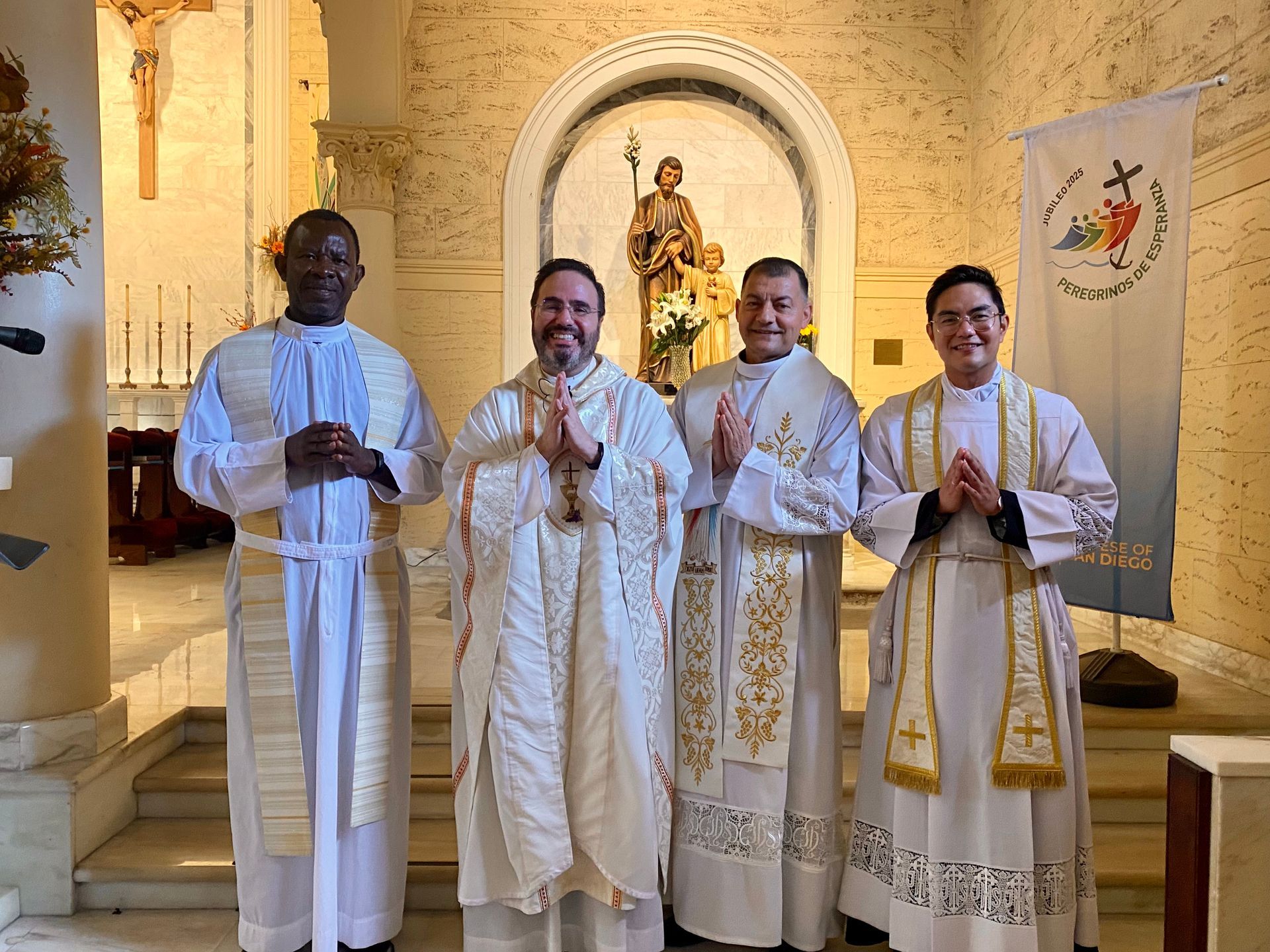 Four priests in white vestments stand in a church, hands clasped before an altar and statue.