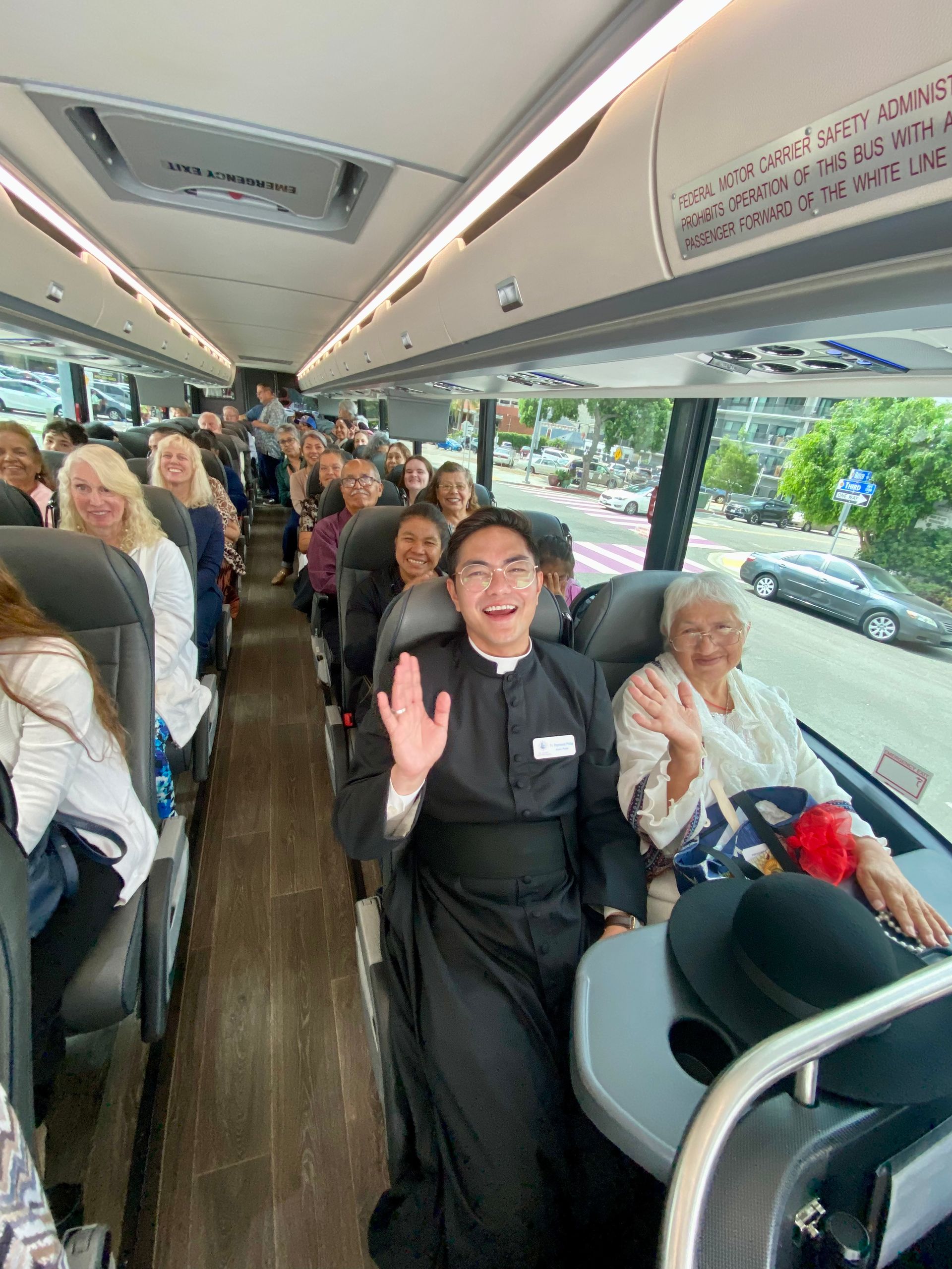 People seated on a bus, smiling and waving inside a bright coach with large windows.
