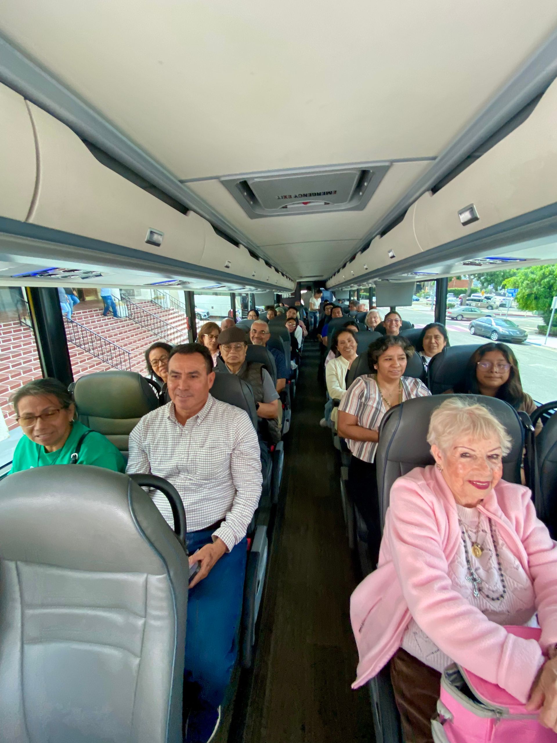 Passengers seated inside a shuttle bus, with an elderly woman in a pink sweater in the foreground
