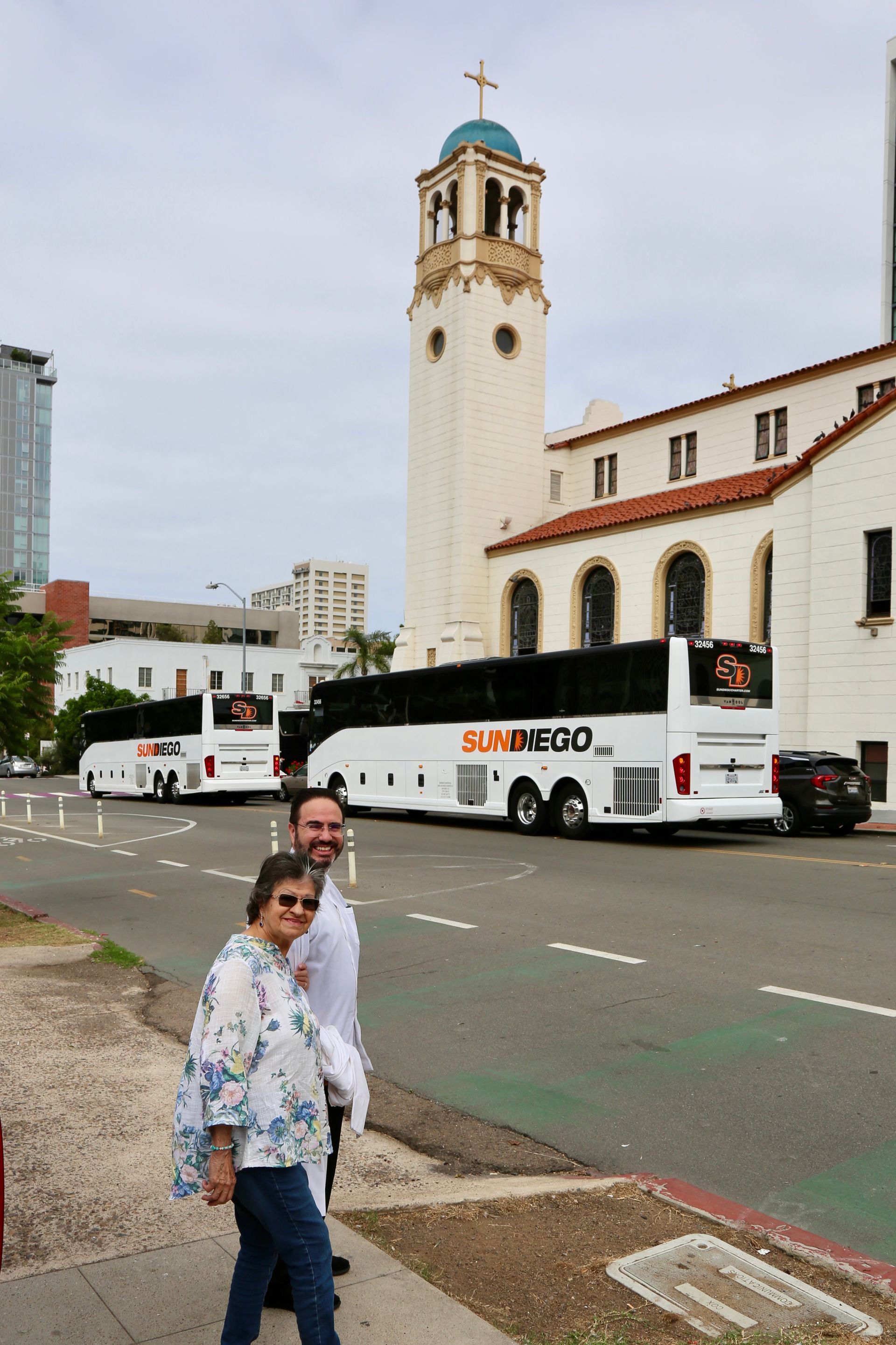 Two people near a white bus and a church tower on a city street