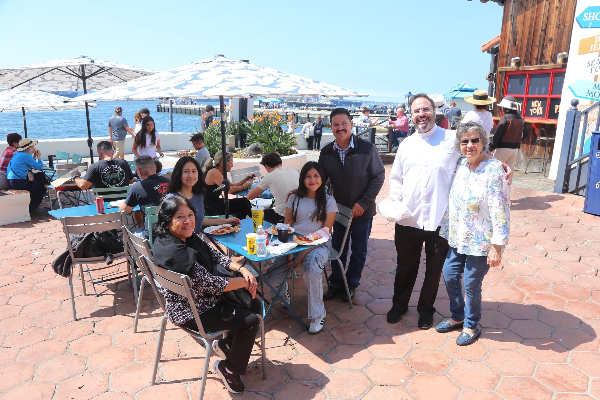 Group of people seated at a seaside café with umbrellas, smiling on a sunny terrace.