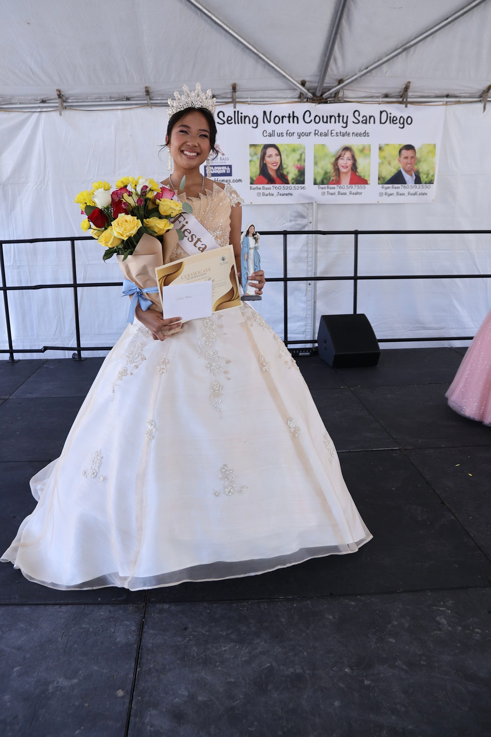 Woman in a white gown holding flowers and trophy on a stage under a tent.