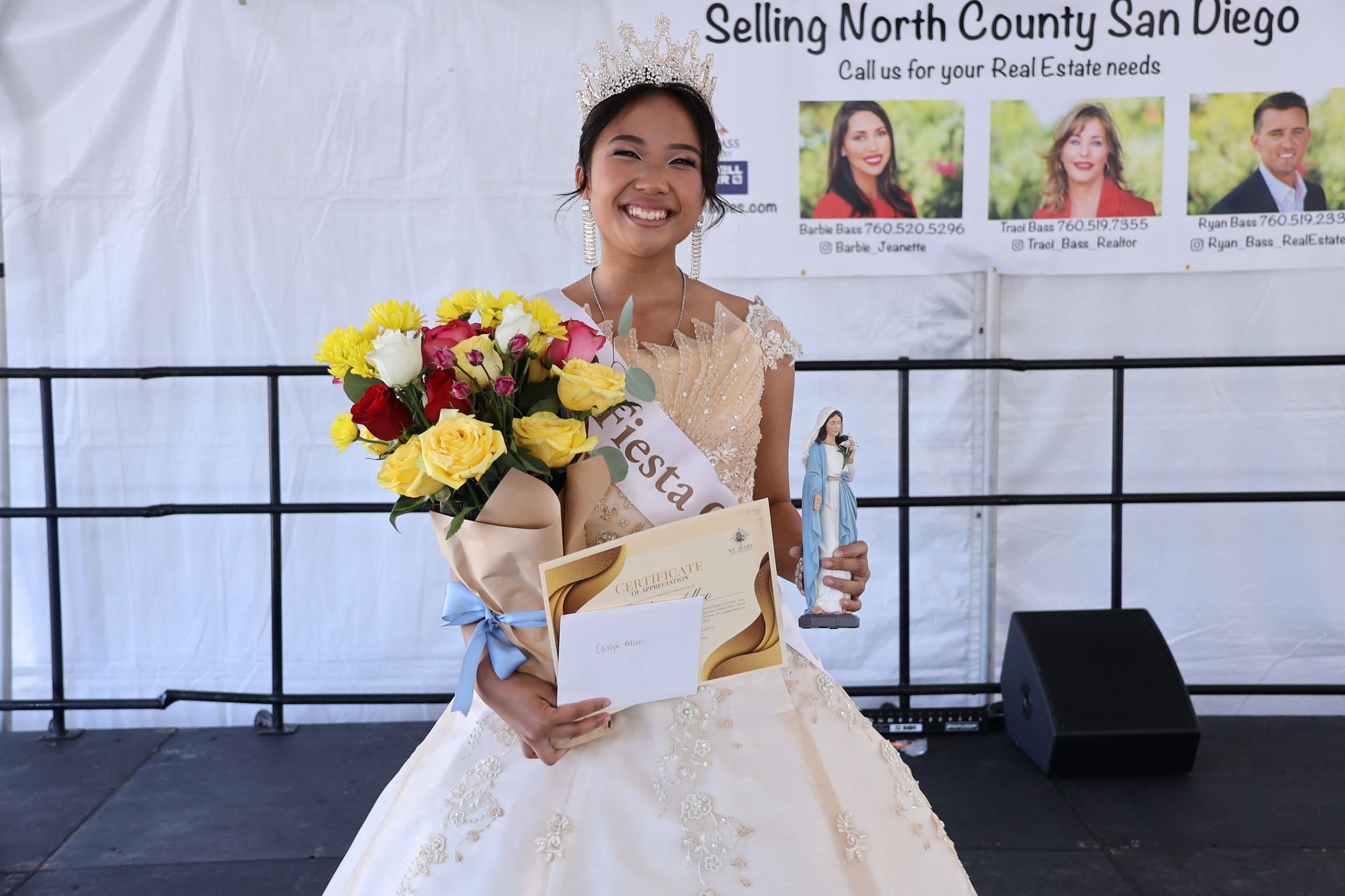 Smiling award recipient in white gown holding flowers and plaque on stage at San Diego event