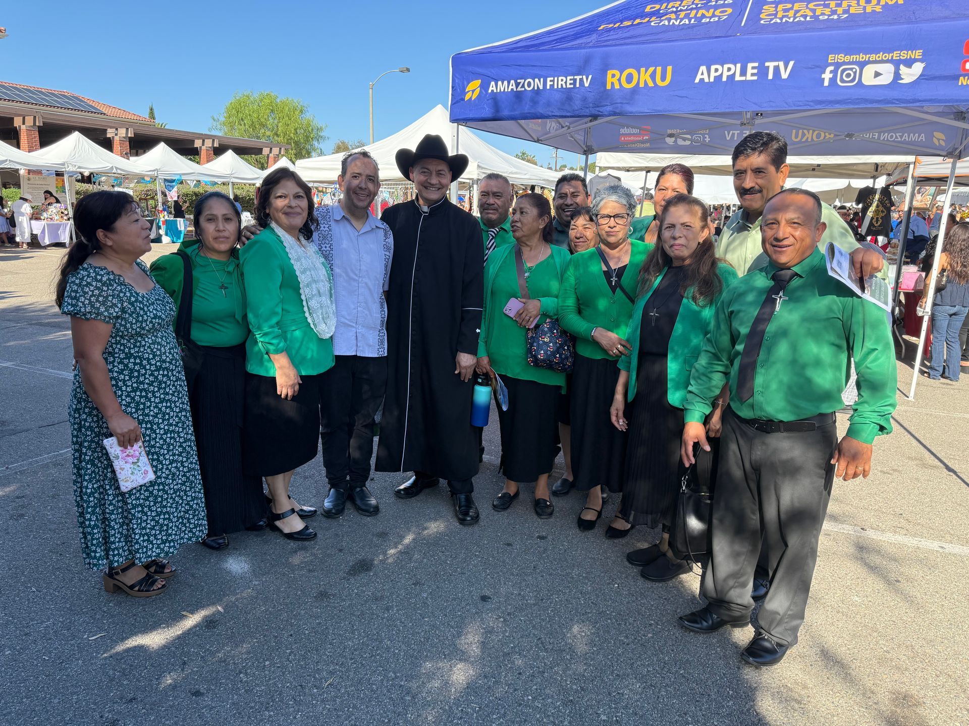Group of people posing outdoors at a festival booth, many wearing green shirts and smiling under a blue canopy.