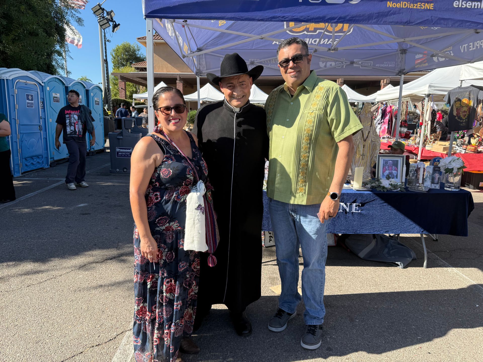 Three people posing at an outdoor market booth in bright sunlight, with tents and vendor tables behind them.