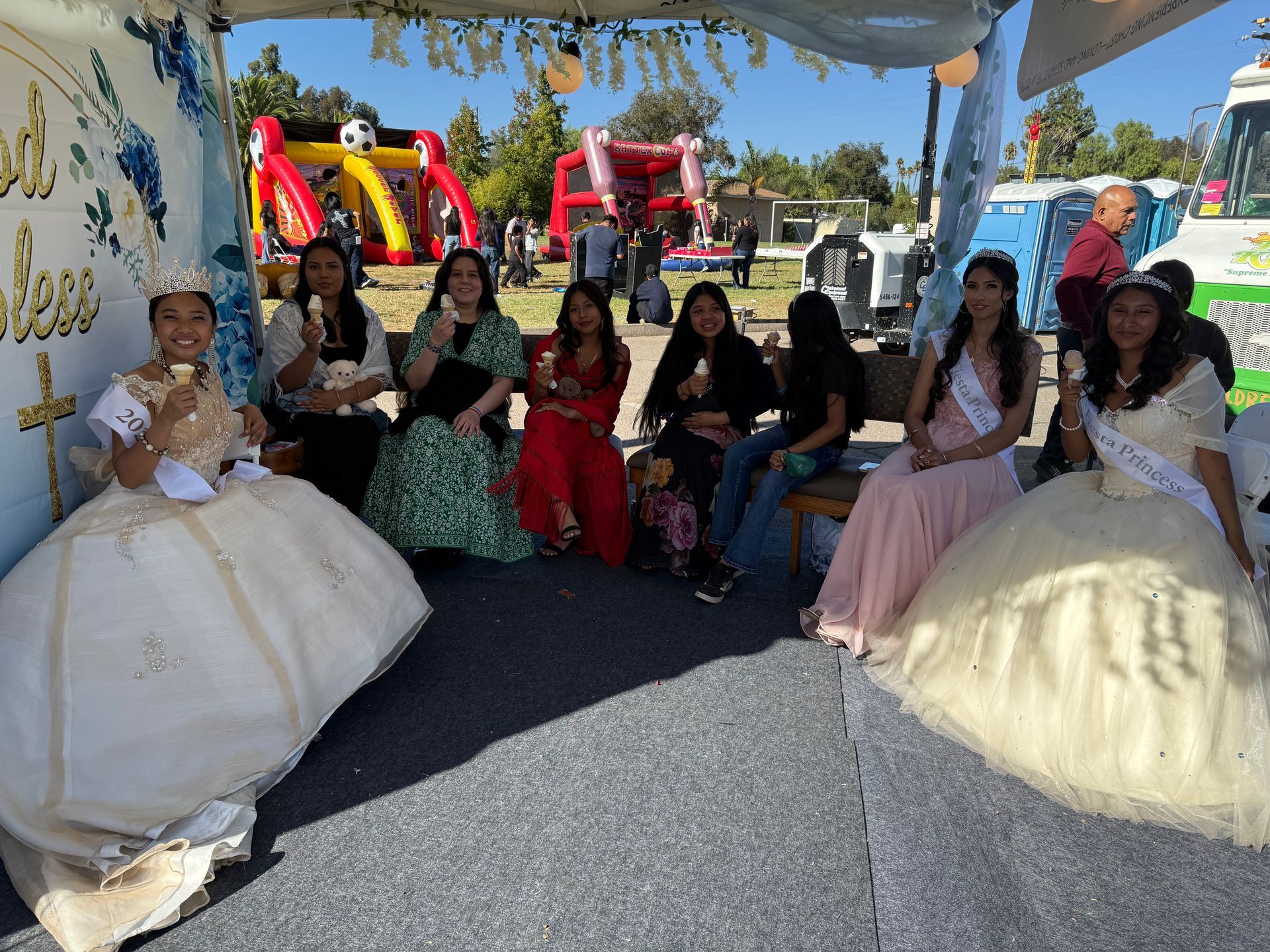 Group seated at an outdoor event under a canopy, wearing colorful formal dresses and smiling at the camera.