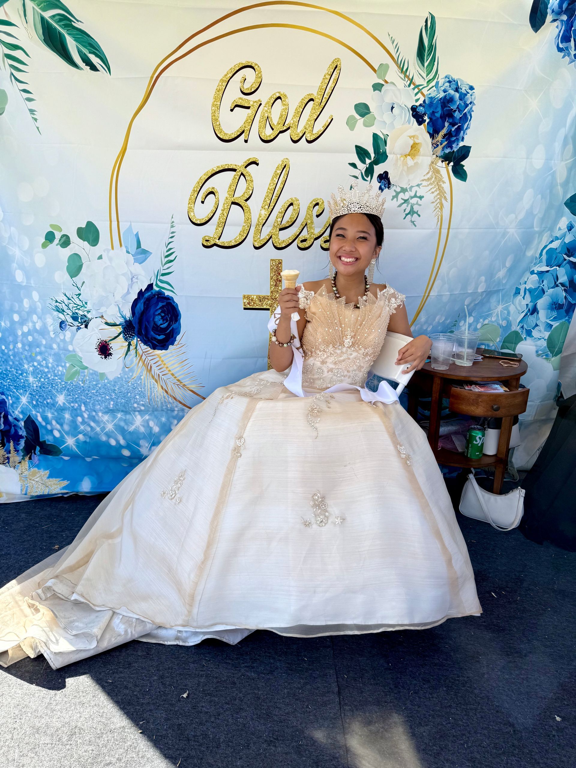 Young woman in a white gown seated before a gold “God Bless” backdrop with blue floral decorations