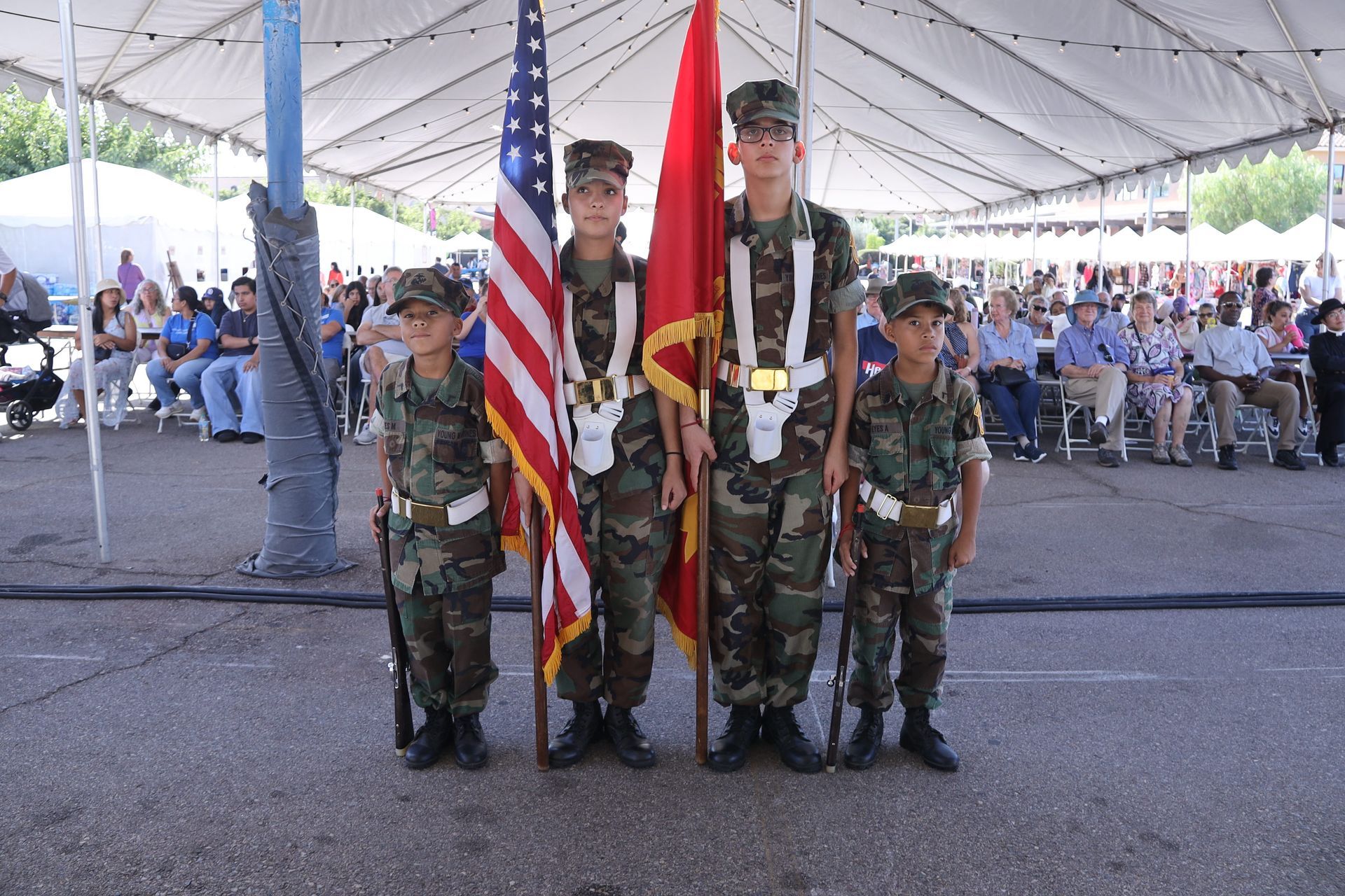 Four uniformed scouts holding flags under a large outdoor event tent with seated audience behind them