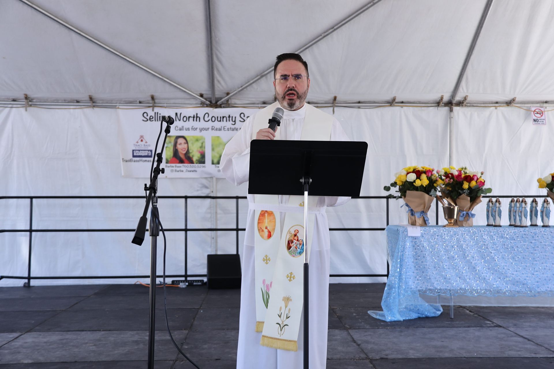 Man speaking at a podium on a stage under a tent, with flowers and awards table beside him