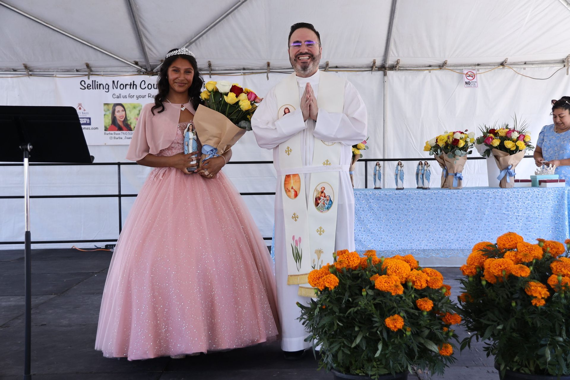 Two people on stage in formal attire, holding bouquets, with orange flowers in front of a white tent backdrop.