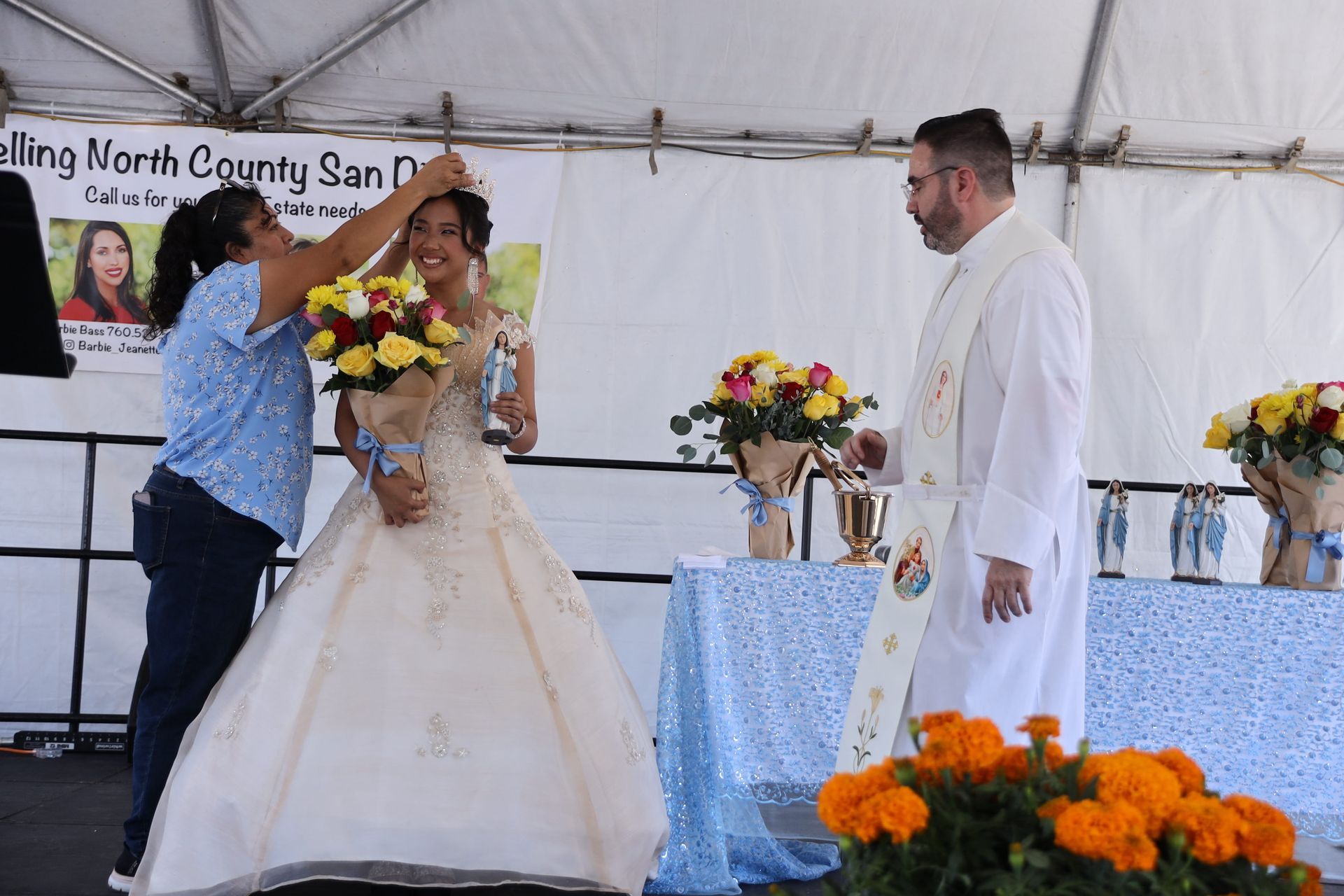 Wedding couple on stage as a woman presents a bouquet; man in white stands nearby under a tent.