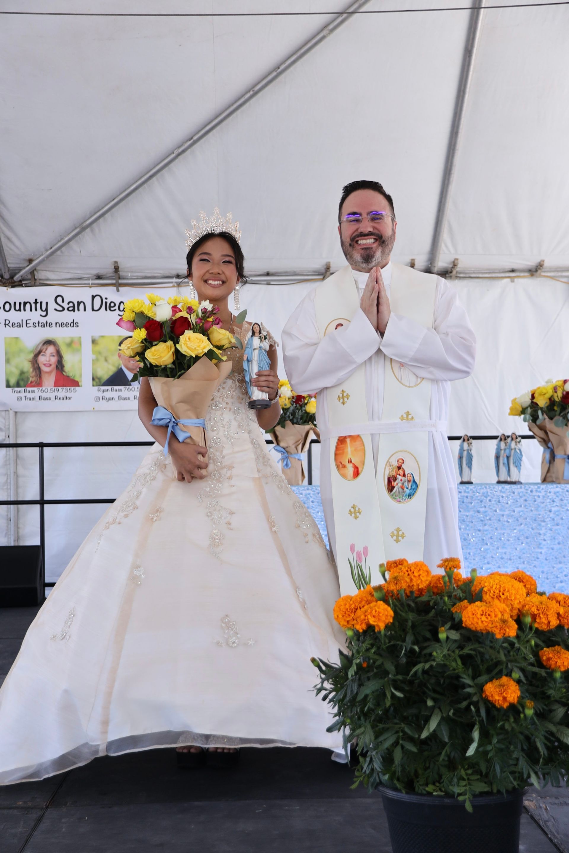 Bride holding flowers and groom in white suit posing on a wedding stage with orange flowers and banner backdrop