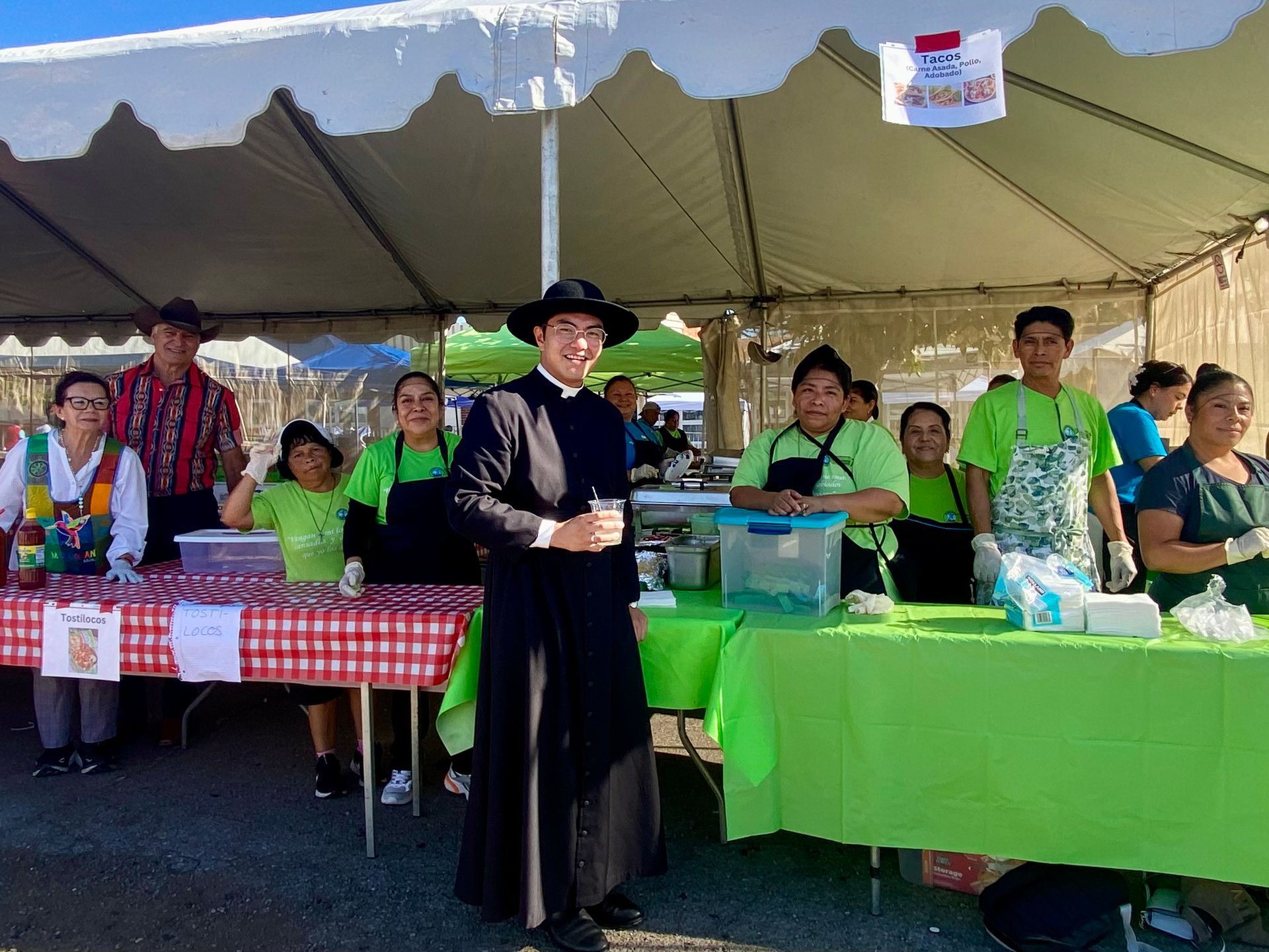 People standing behind market tables under a tent, with a person in black in front and colorful table covers.