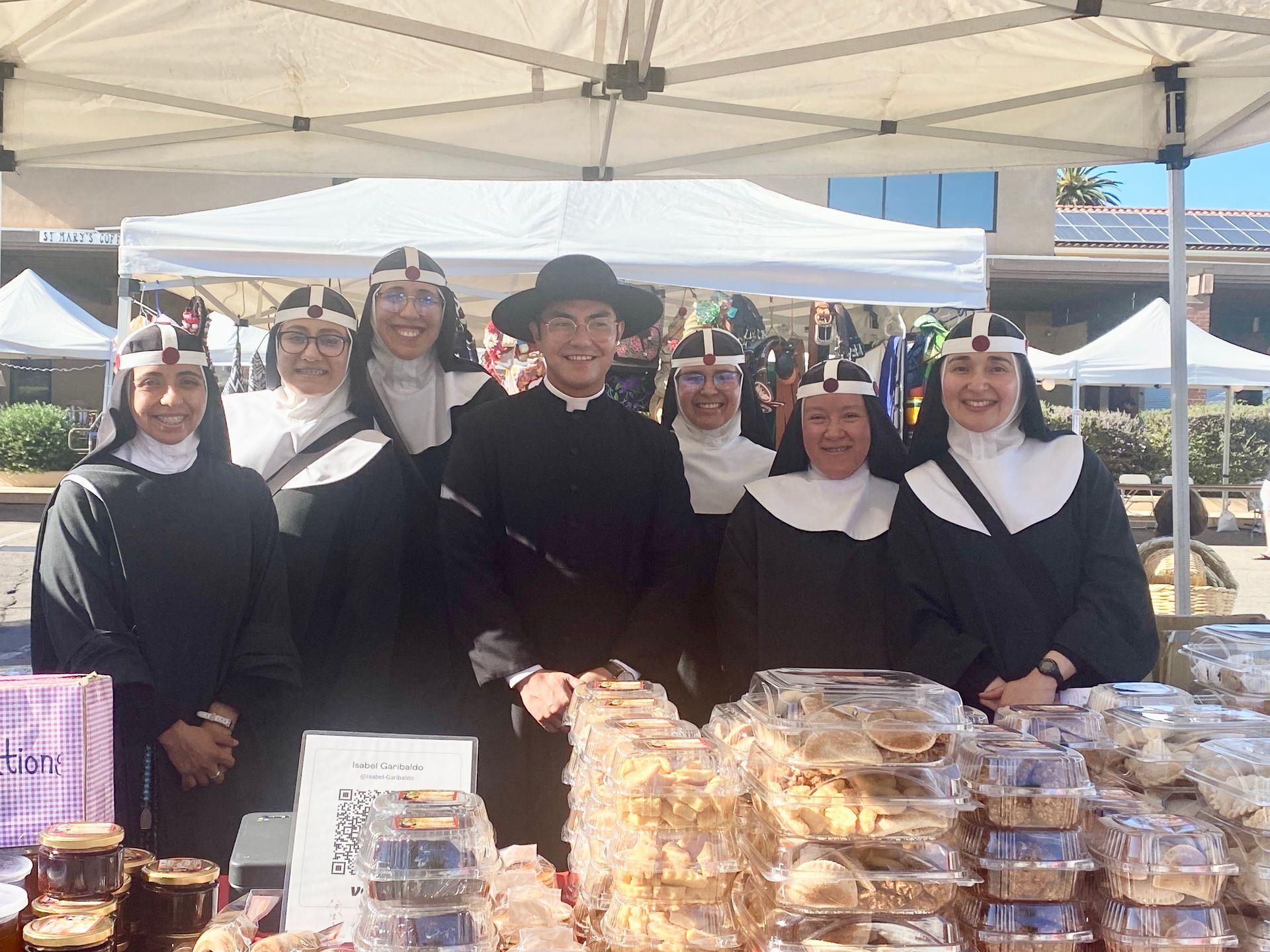 Six people in black-and-white outfits behind a market table stacked with baked goods under a canopy.