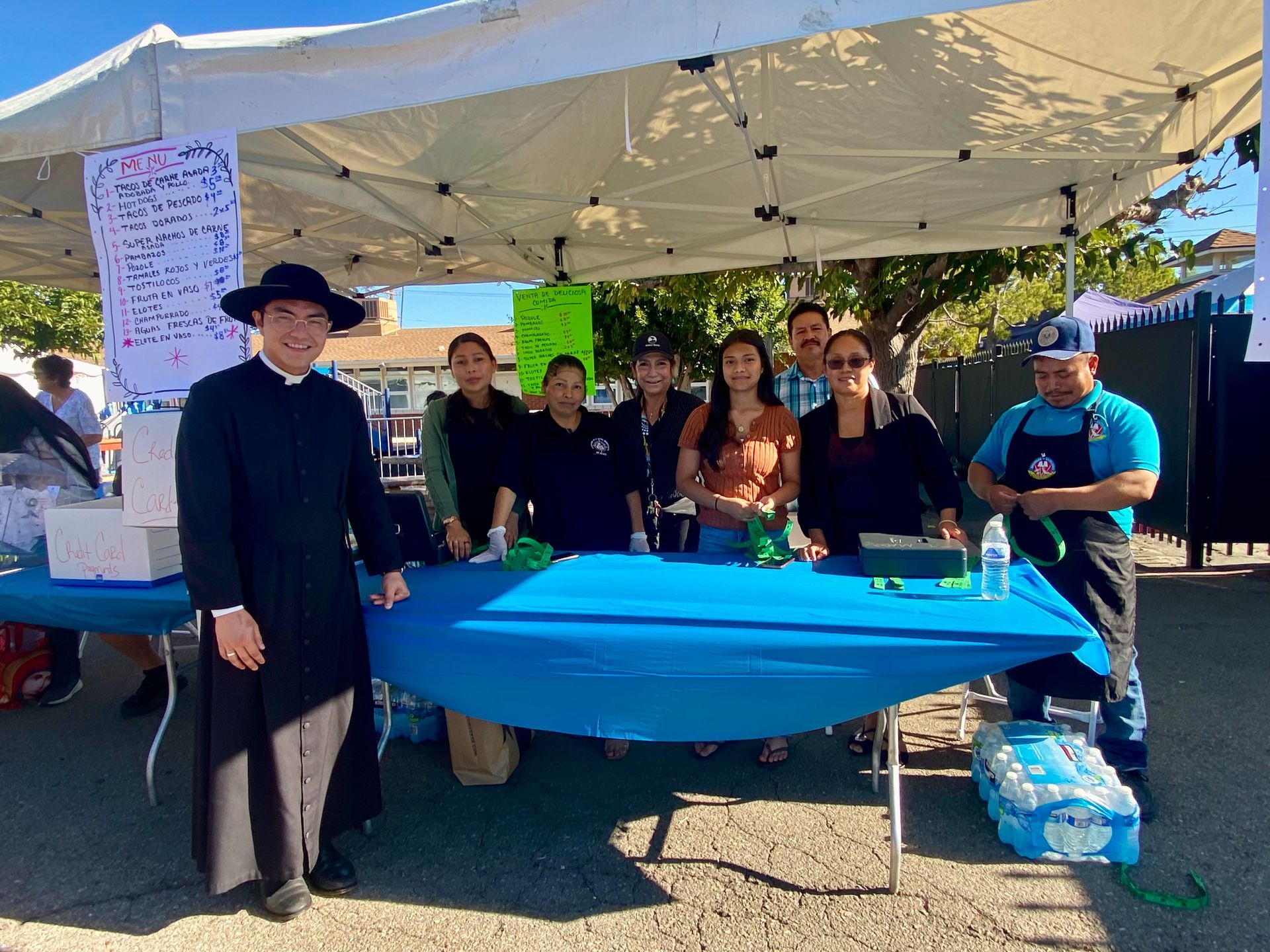 People gathered under a white canopy at an outdoor booth with a blue tablecloth and display materials.