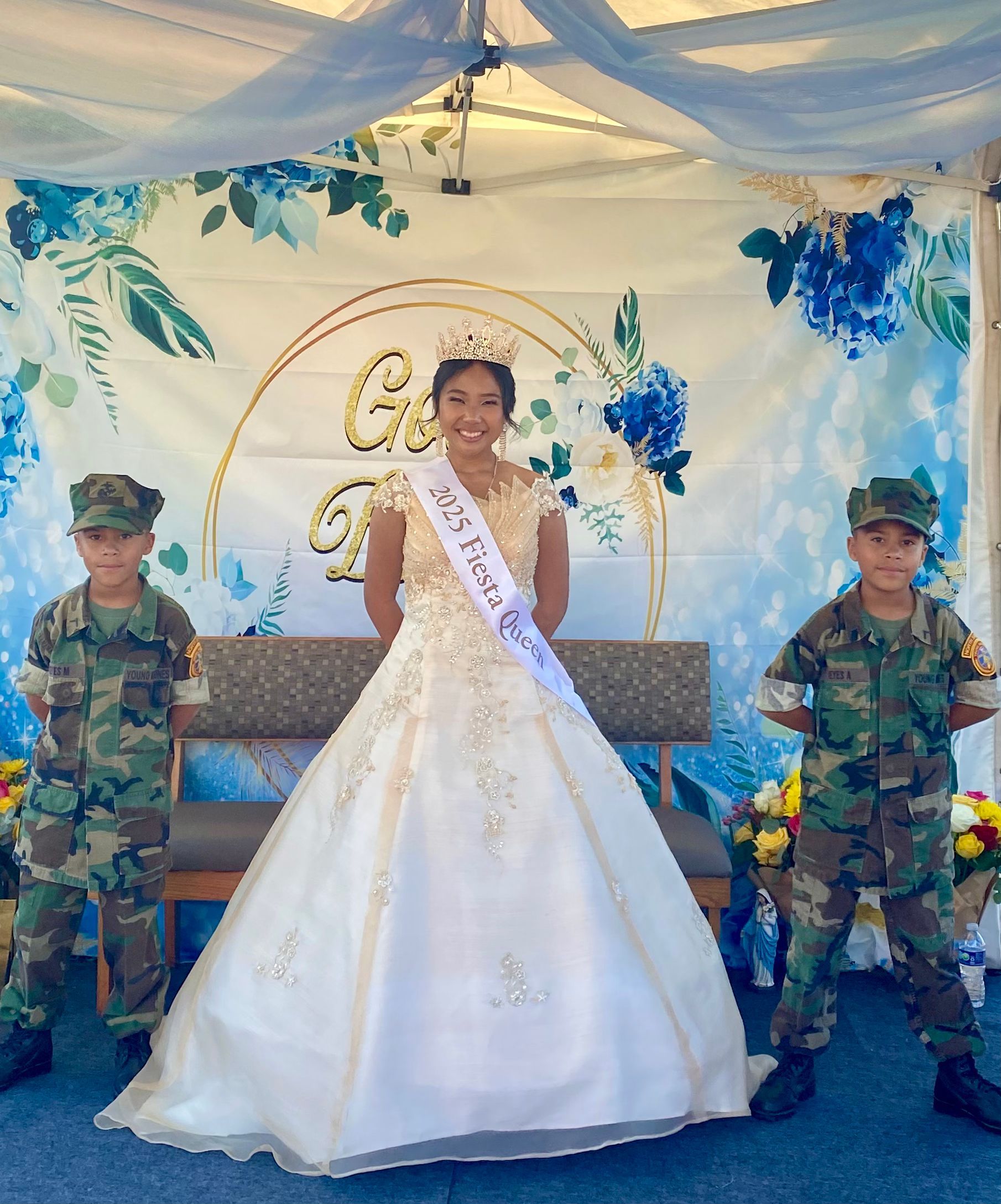 Bride in white gown with sash, standing on a decorated stage beside two uniformed guards.