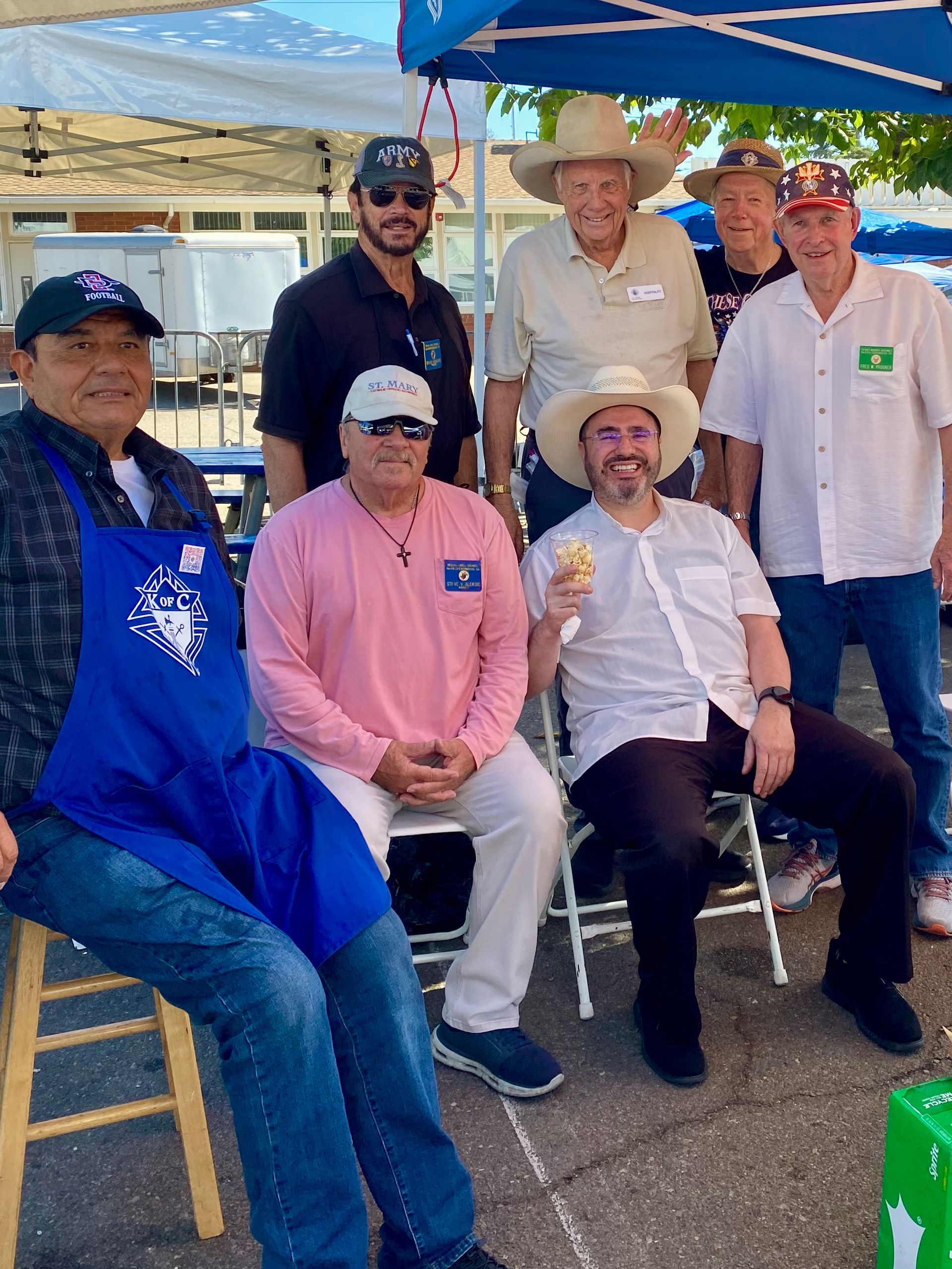Group of six people seated and standing under a blue canopy at an outdoor event, smiling at the camera.