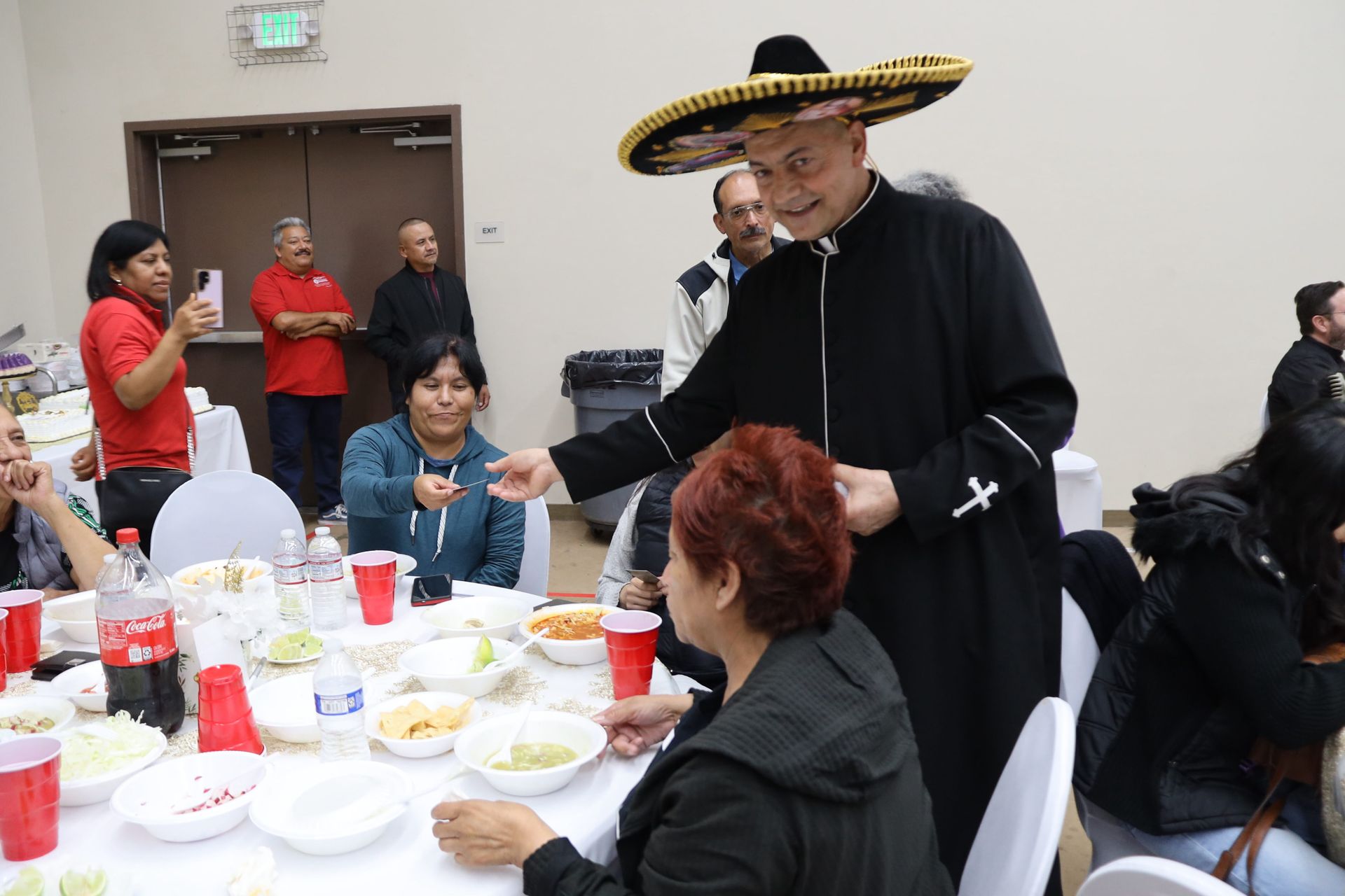 Man in black robe and straw sombrero speaking at a banquet table with seated guests