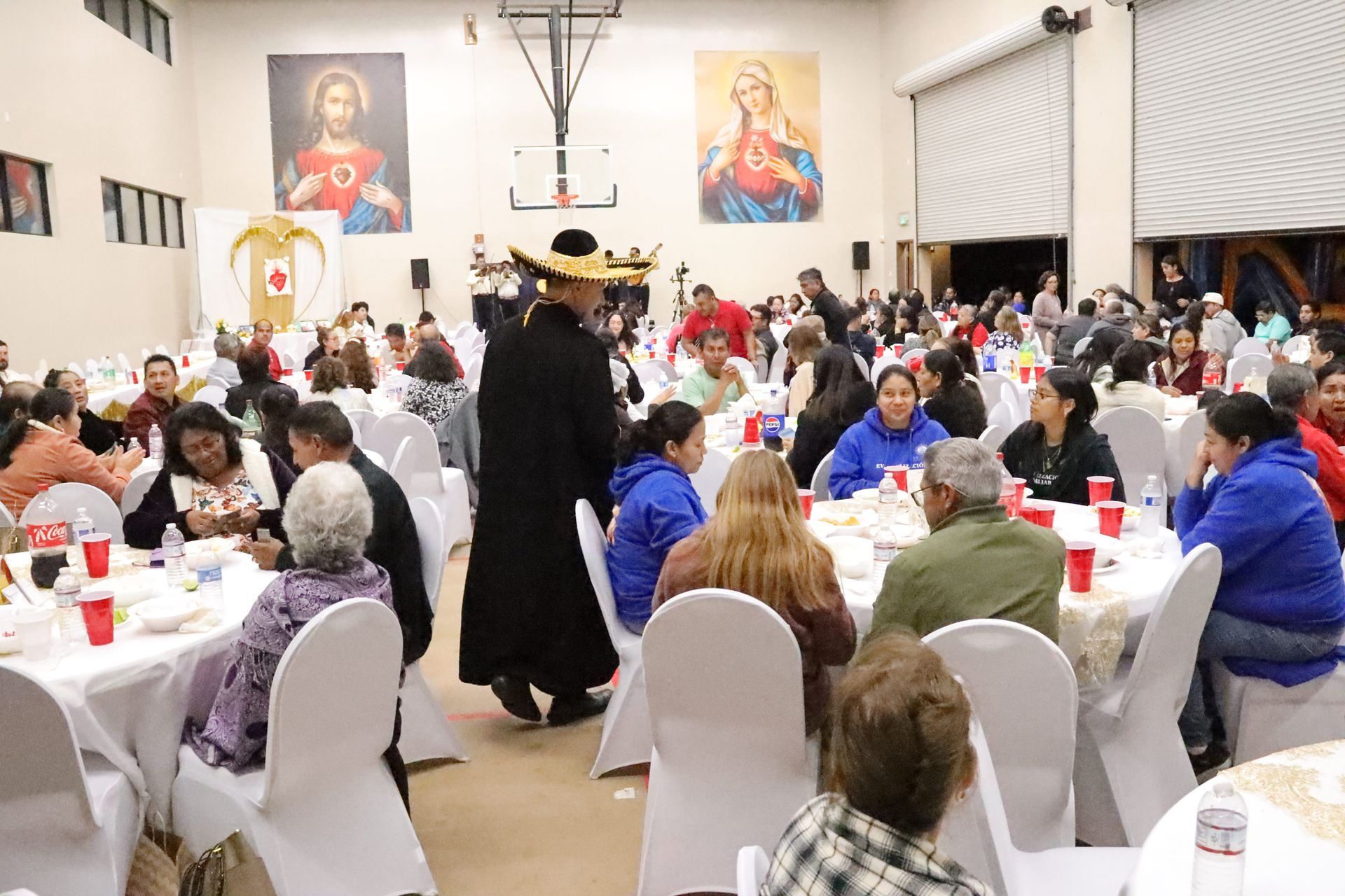 Community banquet hall with guests seated at round tables and a person in black walking between them