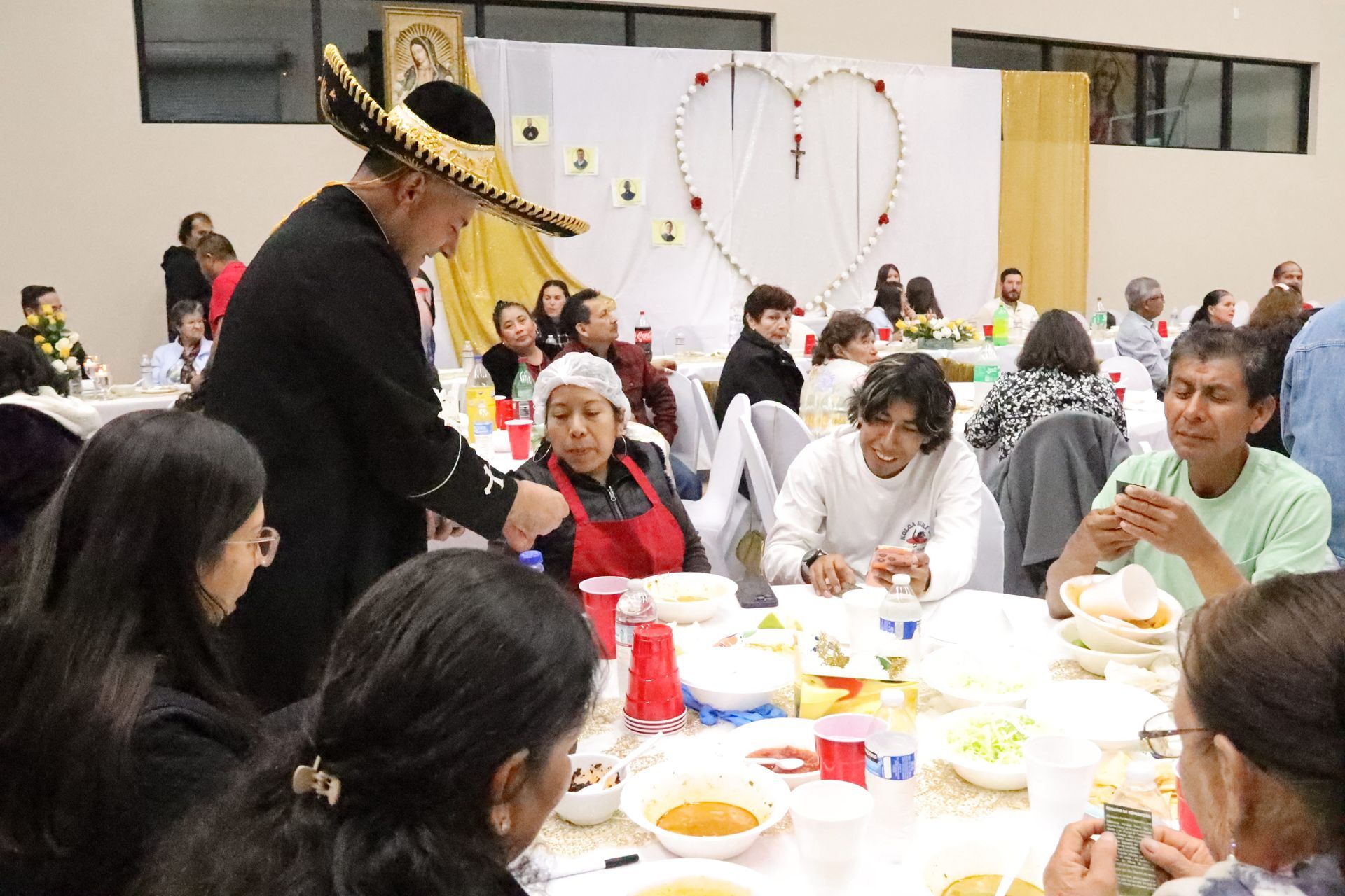 Person in a sombrero serving food at a crowded indoor banquet table with guests seated nearby