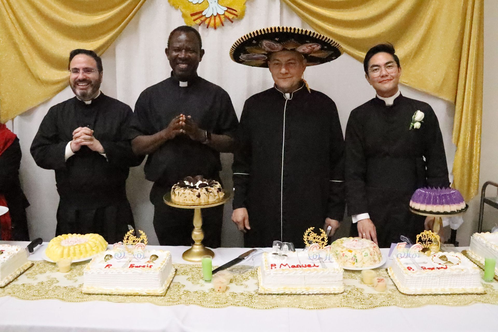 Four men standing behind a decorated table with cakes, smiling at a celebratory indoor gathering