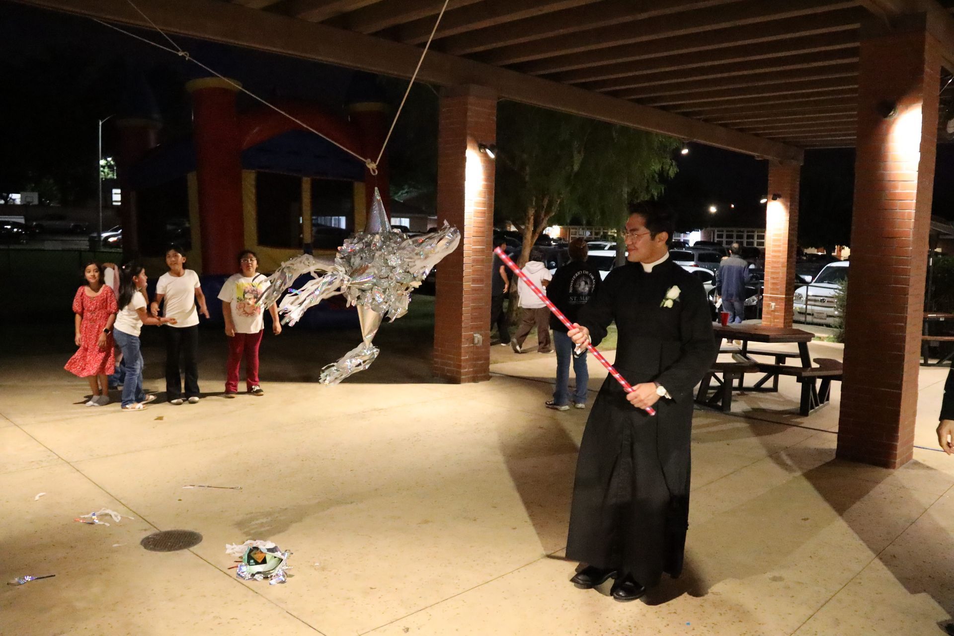 Person in black robe performing with a pink staff under a pavilion as onlookers watch nearby
