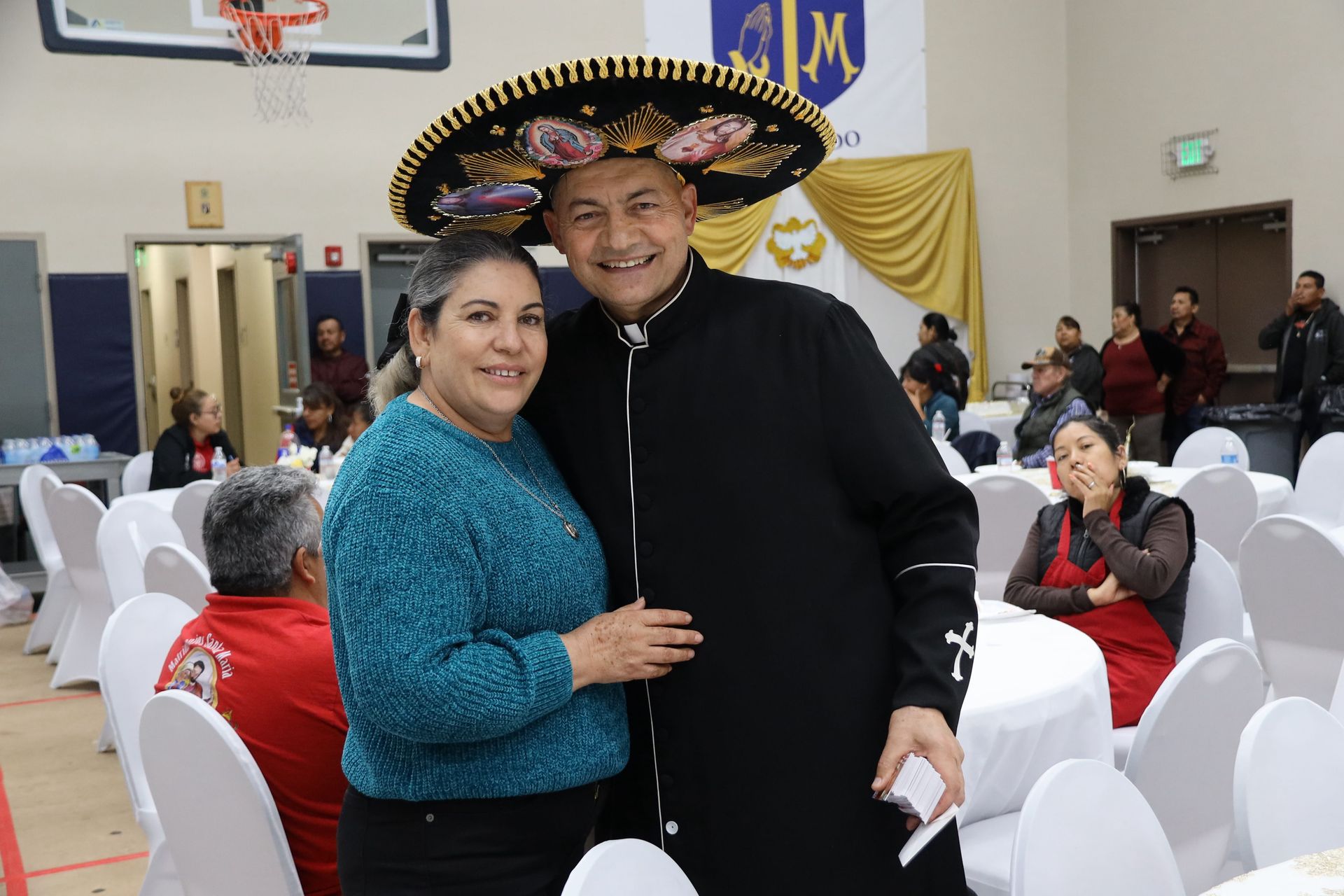 Two smiling people posing together at a festive indoor banquet, one wearing a sombrero