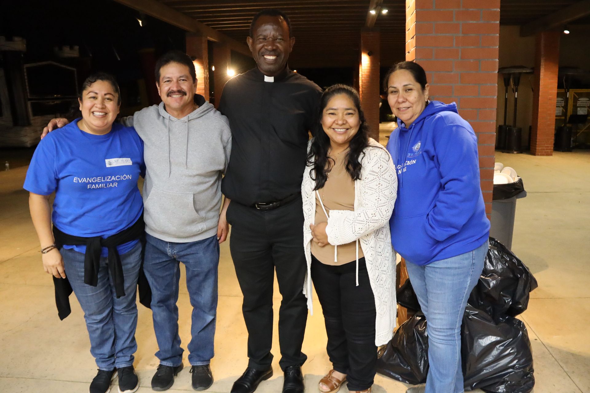 Five people smiling and standing together outdoors at night near a brick building, two holding trash bags