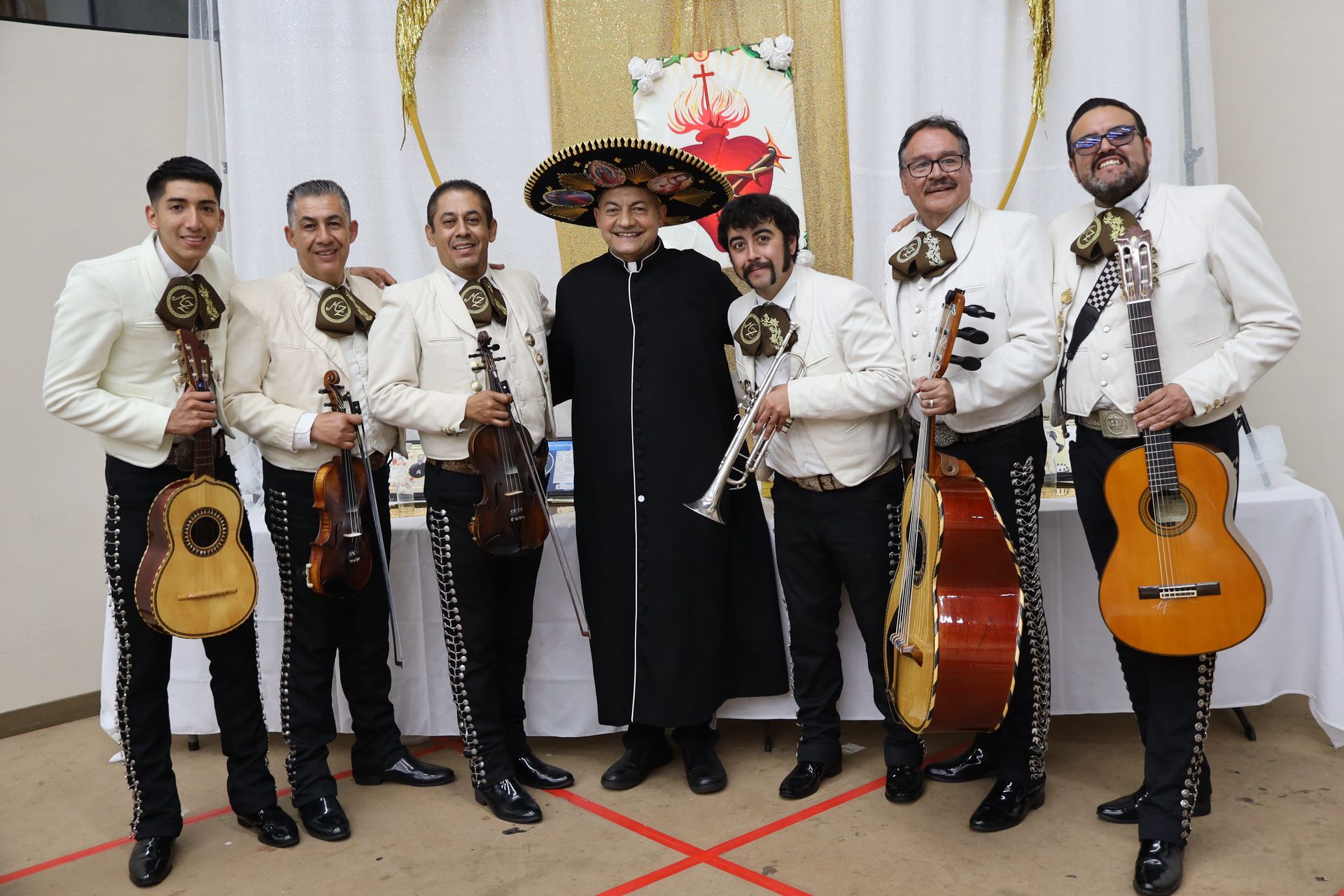 Six musicians in matching white charro outfits pose indoors with guitars and a trumpet.