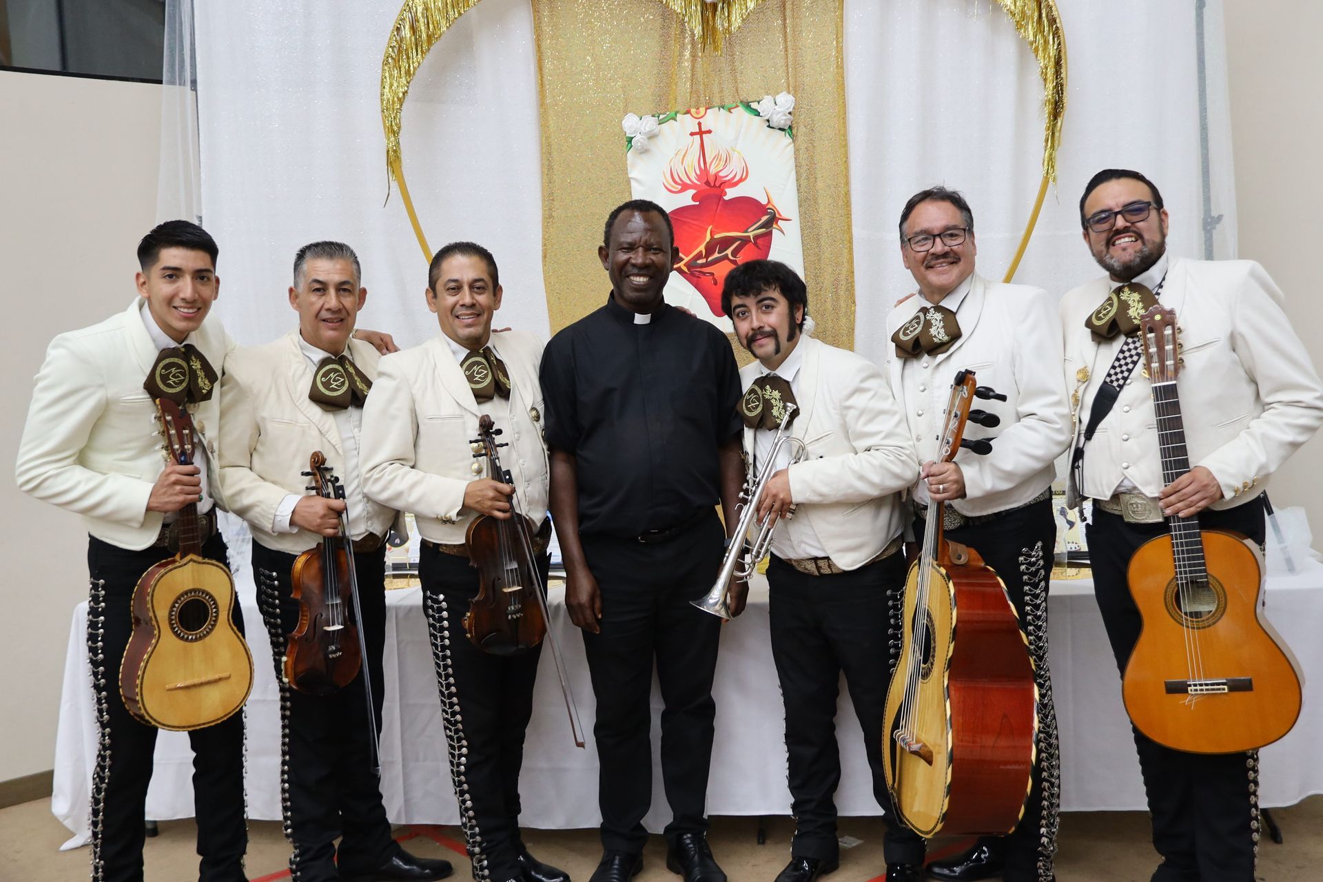 Six musicians in white shirts hold guitars and violins in a decorated indoor event space.