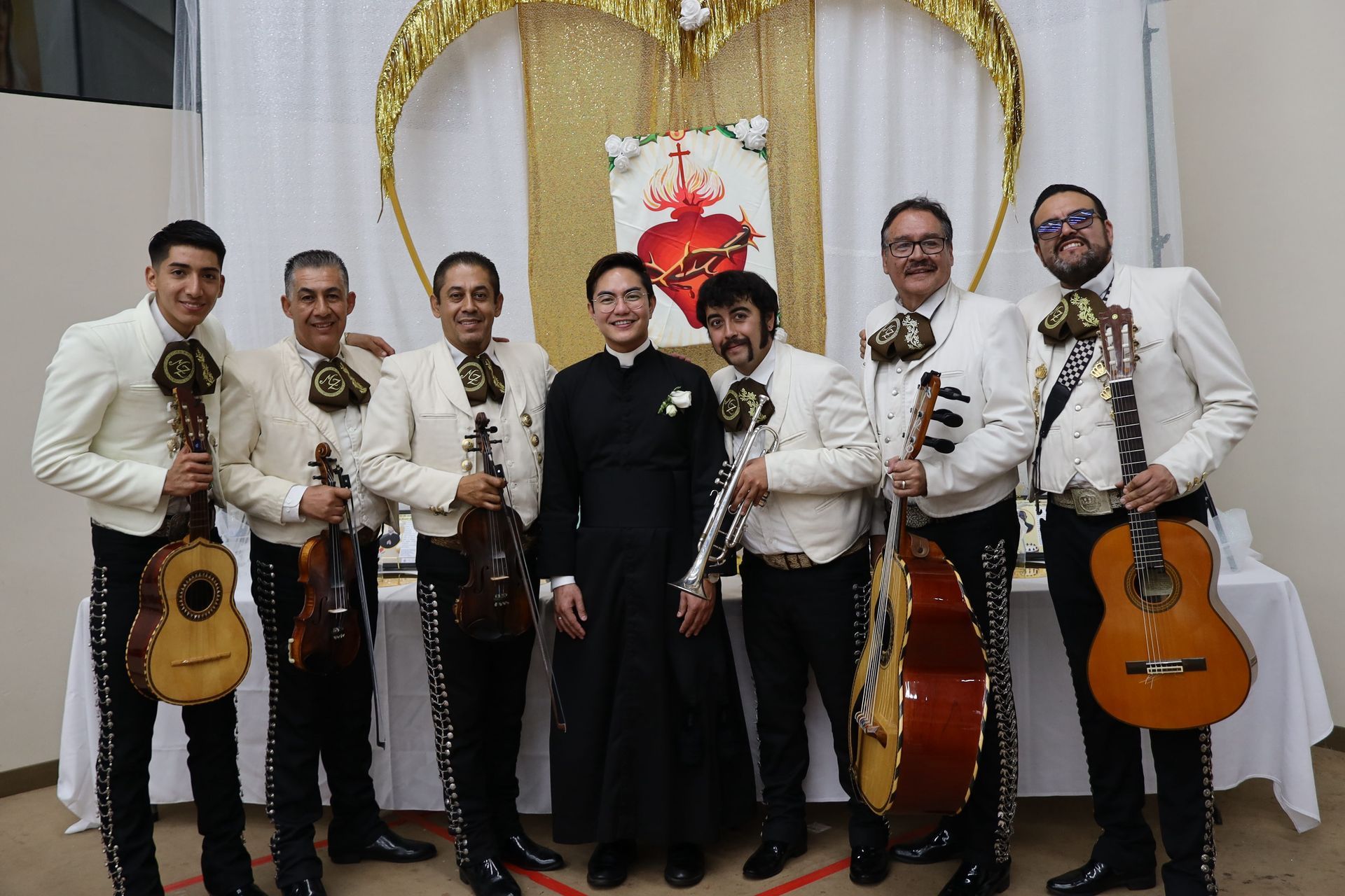 Seven musicians in white shirts pose with guitars and a violin in front of a gold heart backdrop.