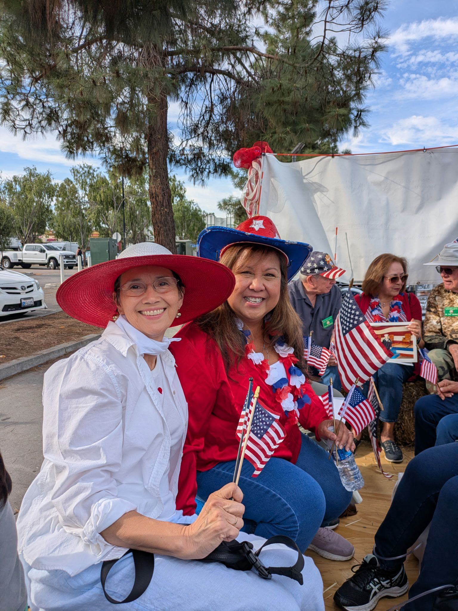 Two people in red, white, and blue hats smile while holding small American flags outdoors.