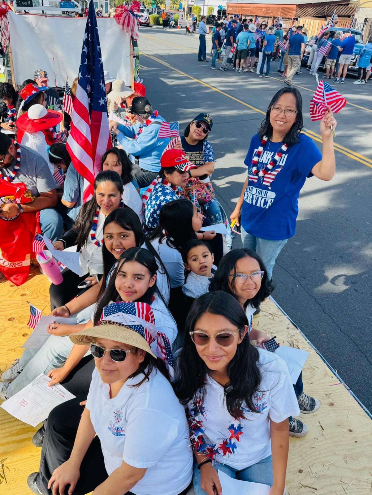 People seated on a parade curb, waving small American flags in red, white, and blue attire