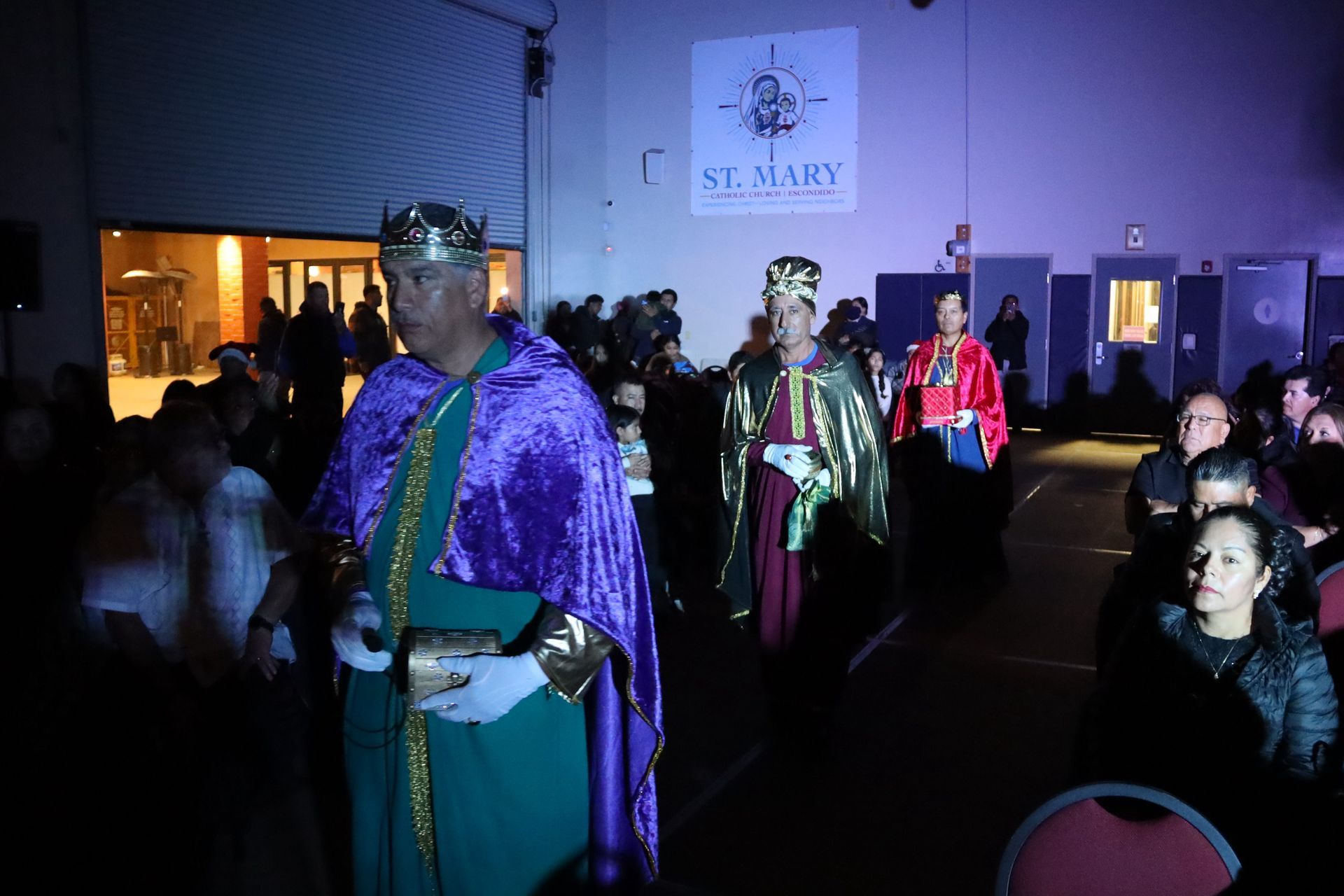 People in colorful costumes and crowns at a nighttime indoor event, with a seated audience watching.