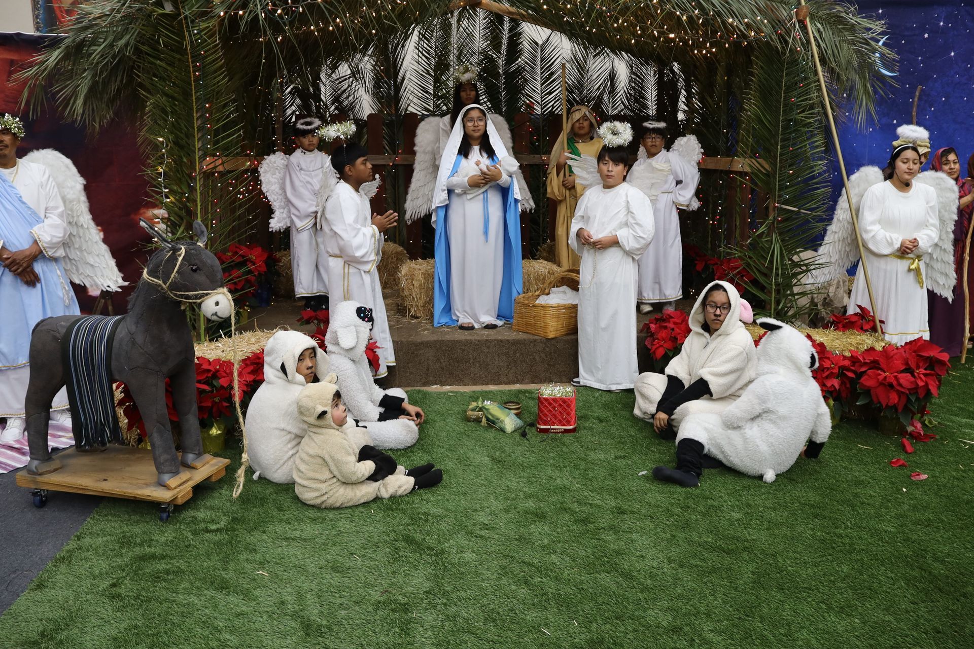 Nativity scene with figures in white and blue robes, a small donkey, and a manger under a thatched canopy