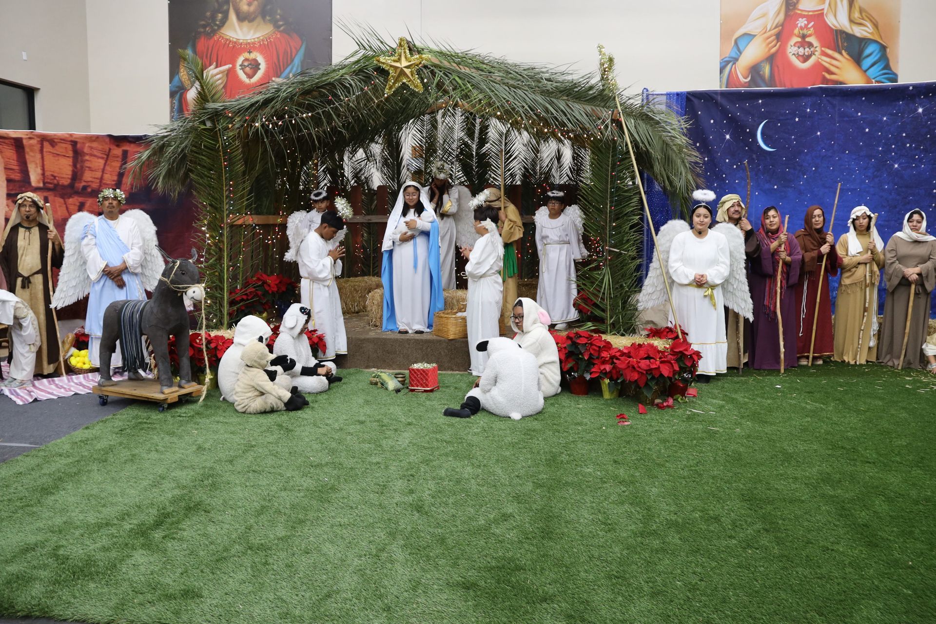 Nativity scene display with people in white robes under a decorated canopy, on green turf with poinsettias