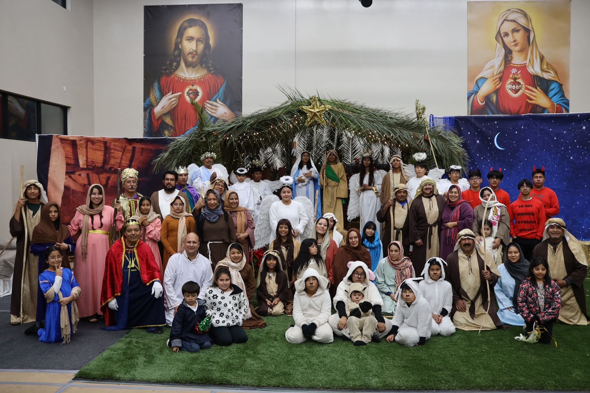 Large group in biblical costumes posing on a stage with nativity decorations and religious murals.
