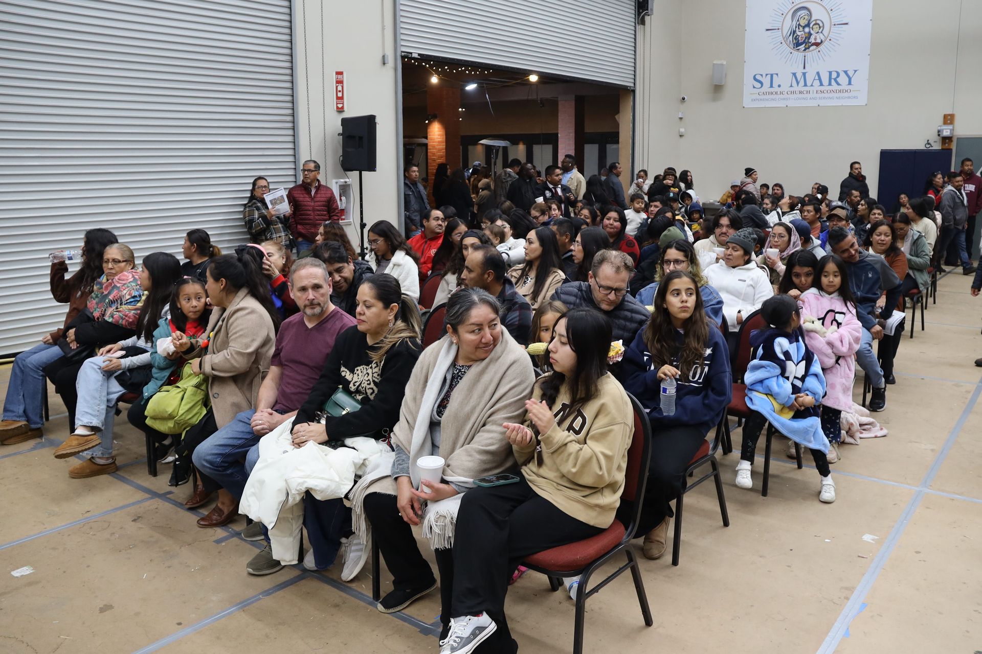 Crowd seated in rows inside a gymnasium at a St. Mary event, with people gathered near the entrance