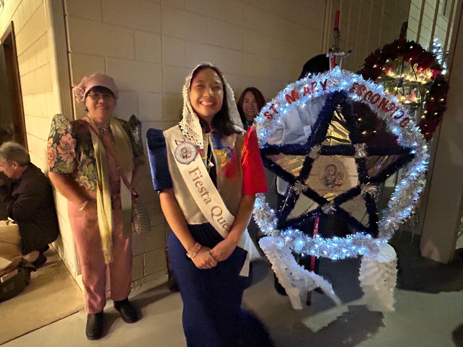 Two people pose beside a glowing holiday decoration in a festive indoor setting.