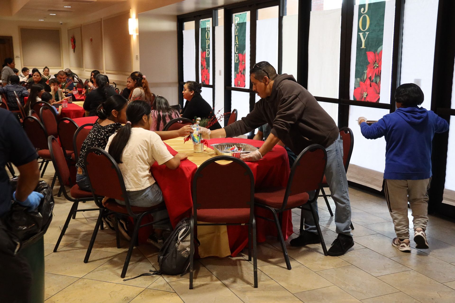 People gathered around tables in a bright hall, with a man leaning over a red table near a window.