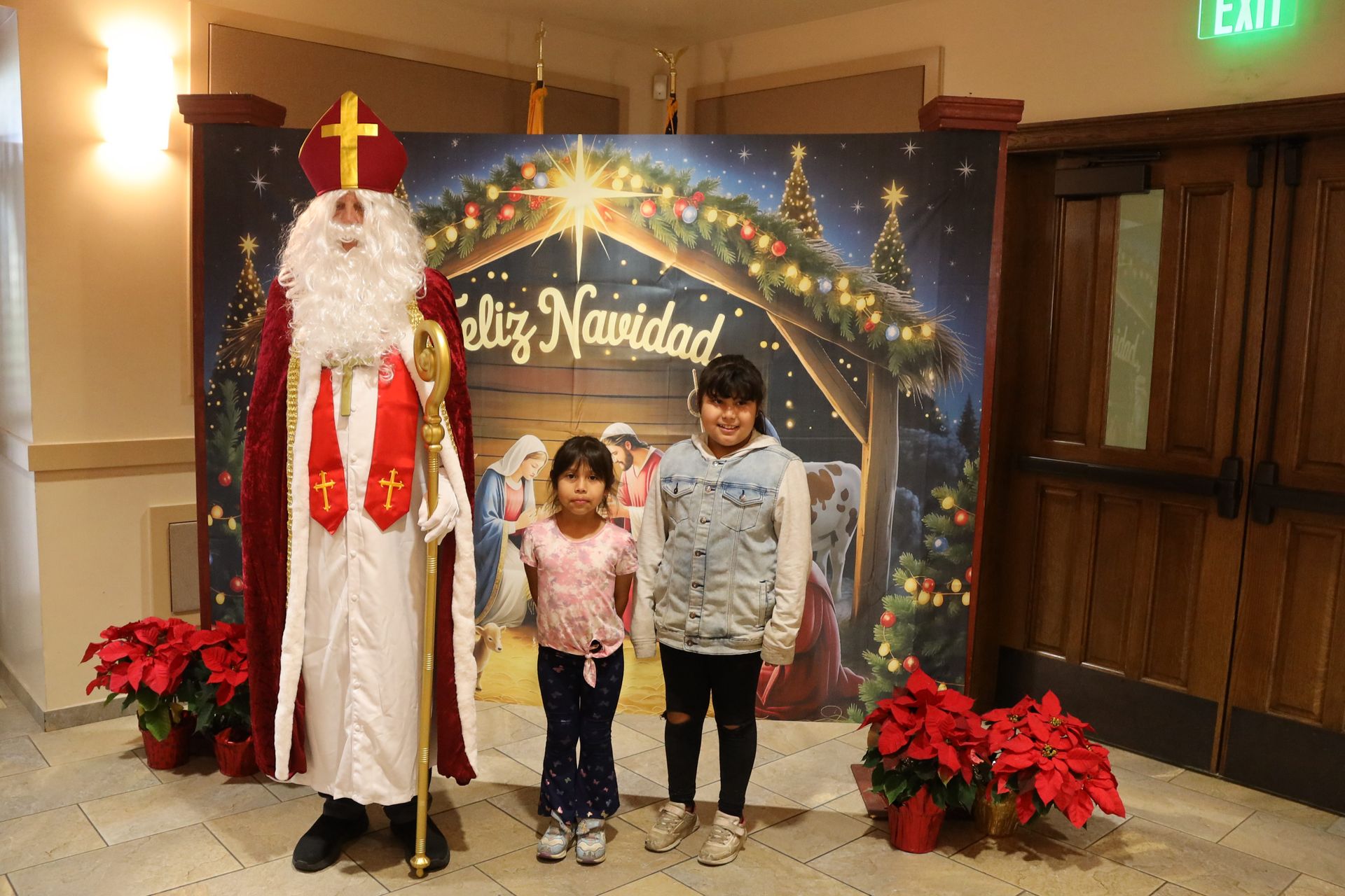 Santa and two children posing in front of a festive Christmas backdrop with poinsettias and an EXIT sign nearby
