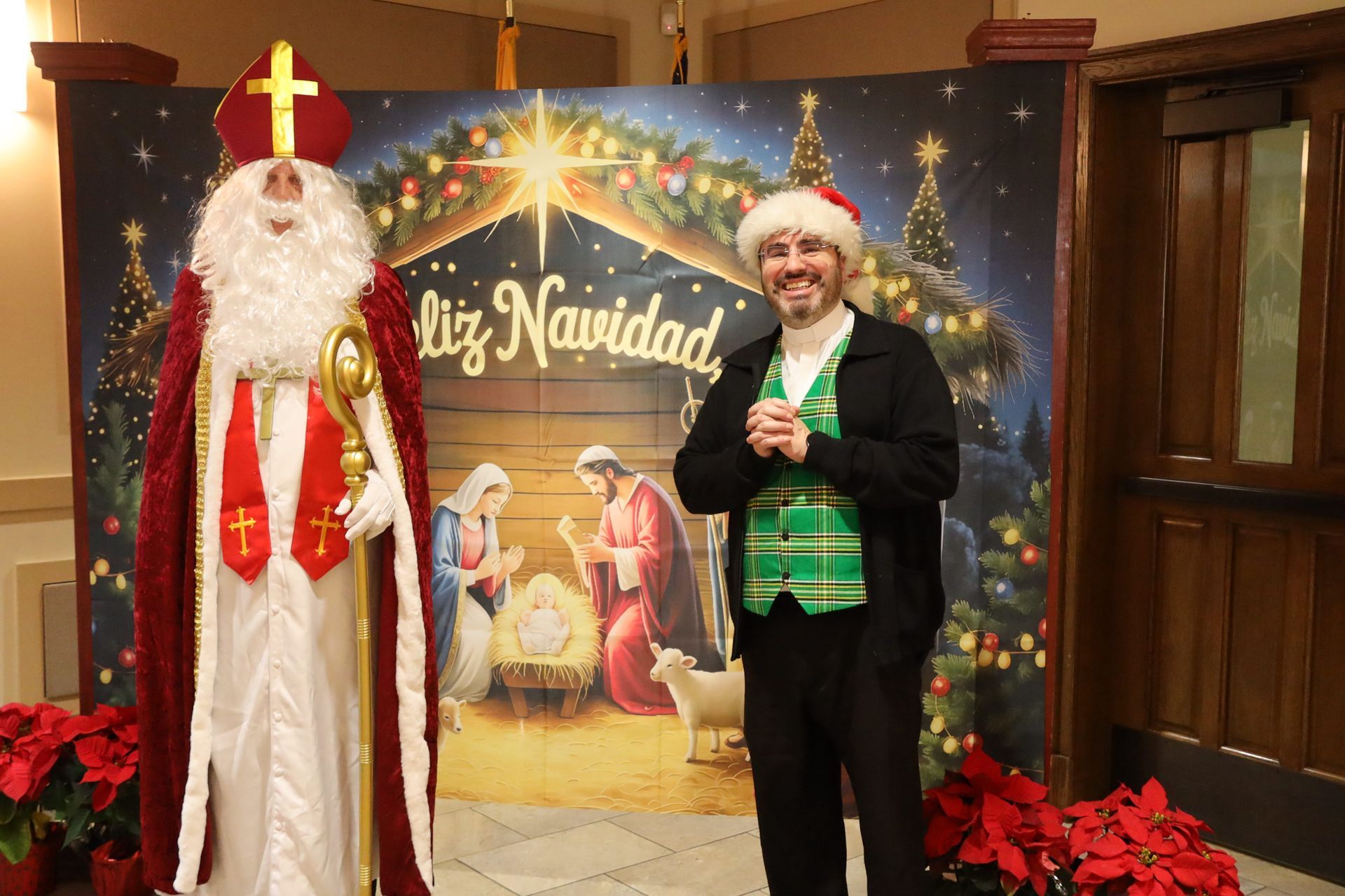 Two people in Santa hats pose before a “Feliz Navidad” holiday display with poinsettias.