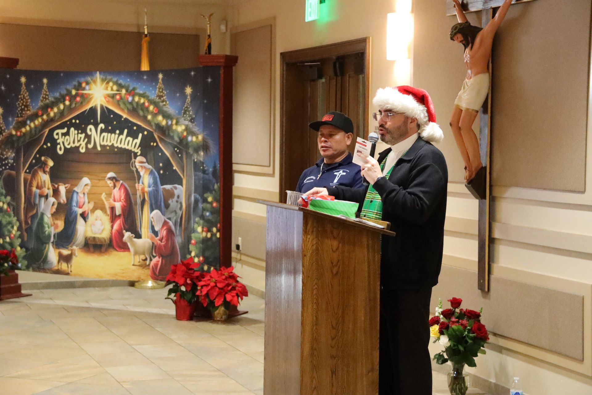 Two men at a podium in Santa hats beside a holiday display and poinsettias indoors